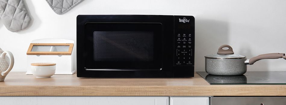 Lifestyle image of black Total Chef microwave on a light-colored wood countertop in front of a white wall. There is a gray oven mitt and potholder hanging on the wall to the left and a black cooktop with a gray saucepan to the right