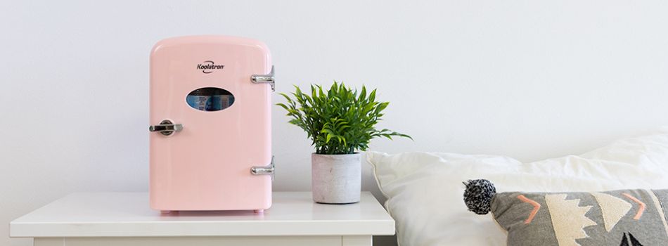 Lifestyle image of a light pink 4L mini fridge on a small white nightstand with a potted plant beside it