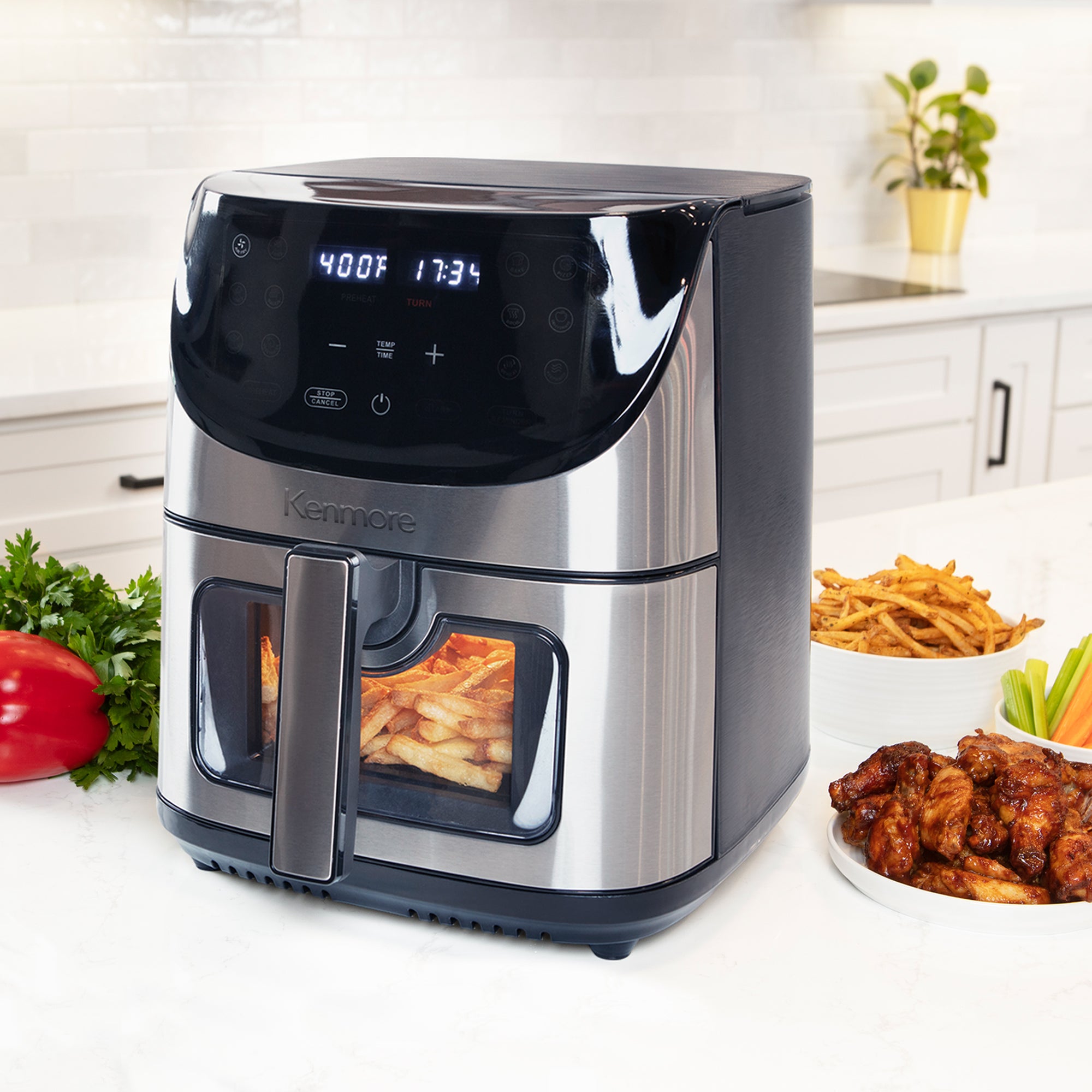 Kenmore digital air fryer on a white kitchen counter with plates of cooked food arranged around it and white cupboards and white tile backsplash in the background.