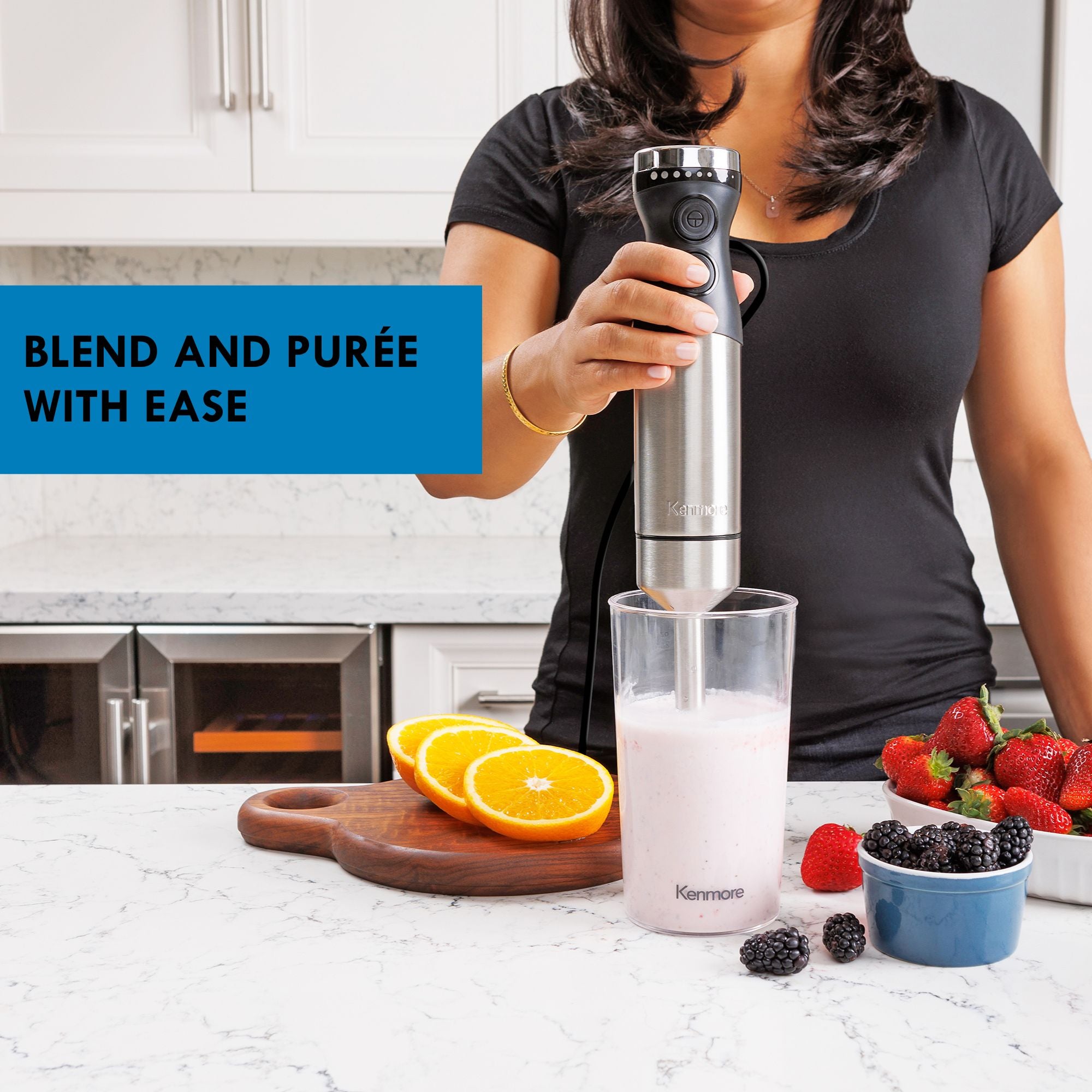 A person in a black t-shirt using the Kenmore hand blender to blend light pink smoothie on a white marble counter with fruits and berries on either side. Text overlay reads, "Blend and puree with ease"