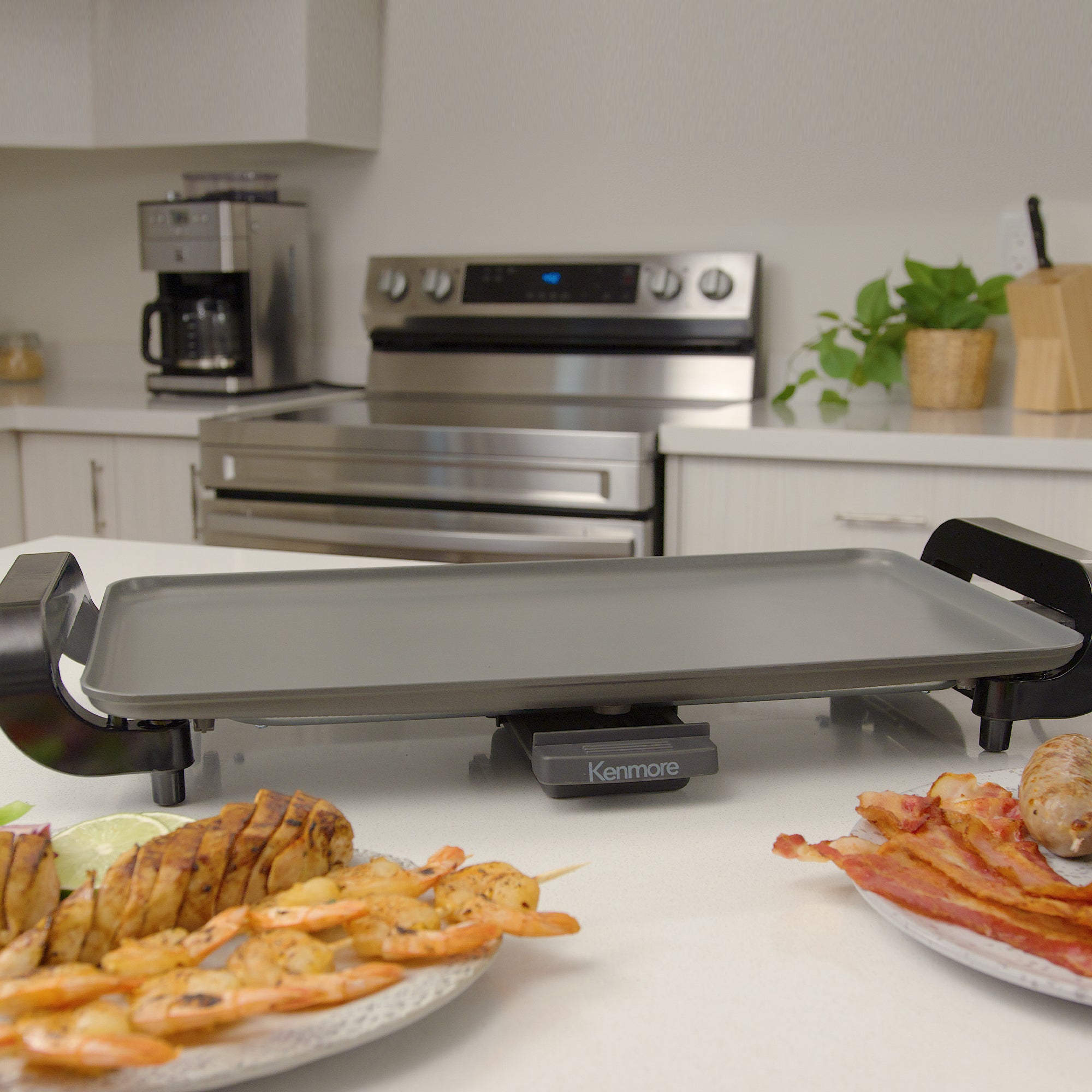 Kenmore non-stick electric griddle on a white countertop with stainless steel appliances in the background and plates of cooked food in front of it
