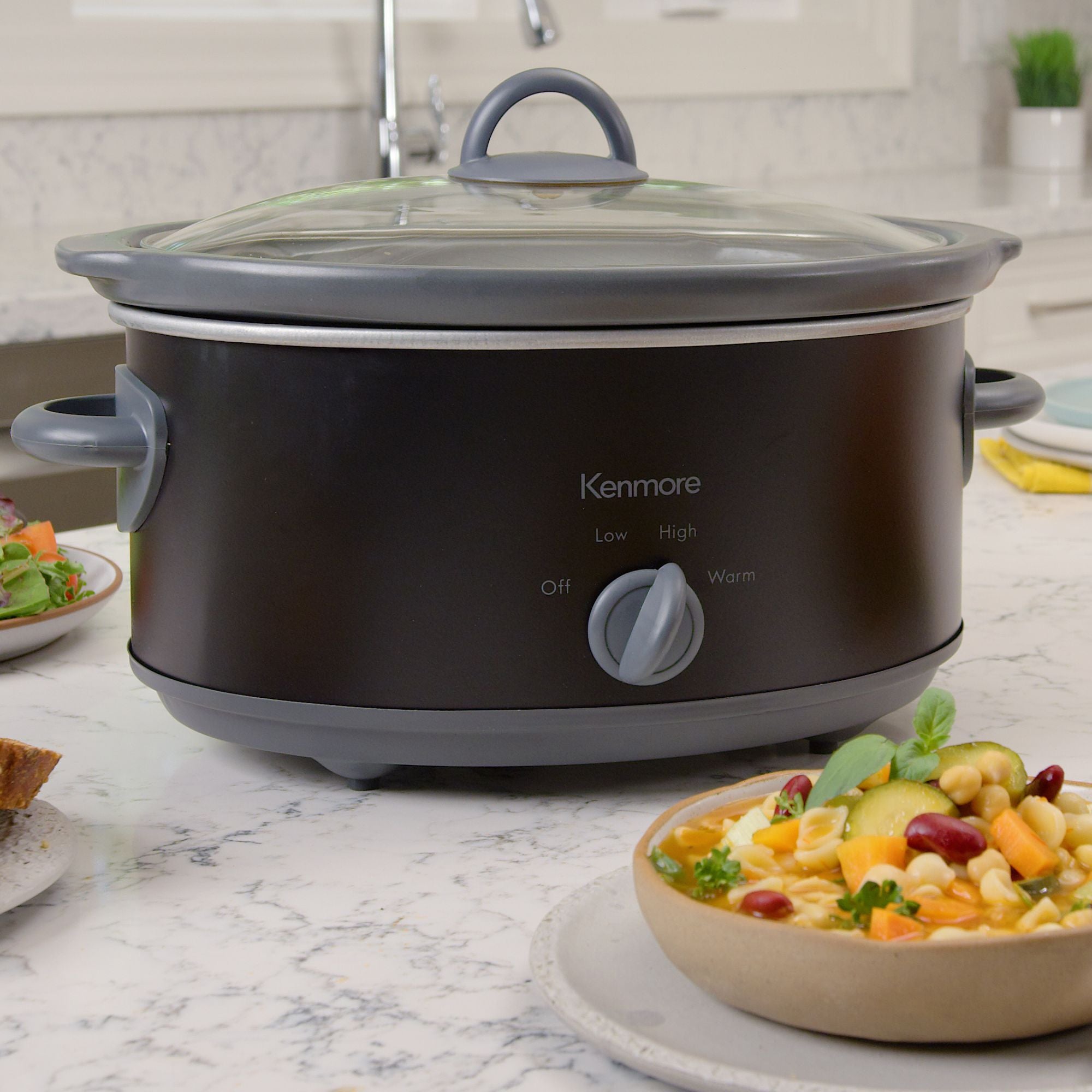 Kenmore 5 qt slow cooker on a white marble countertop surrounded by plates of food 