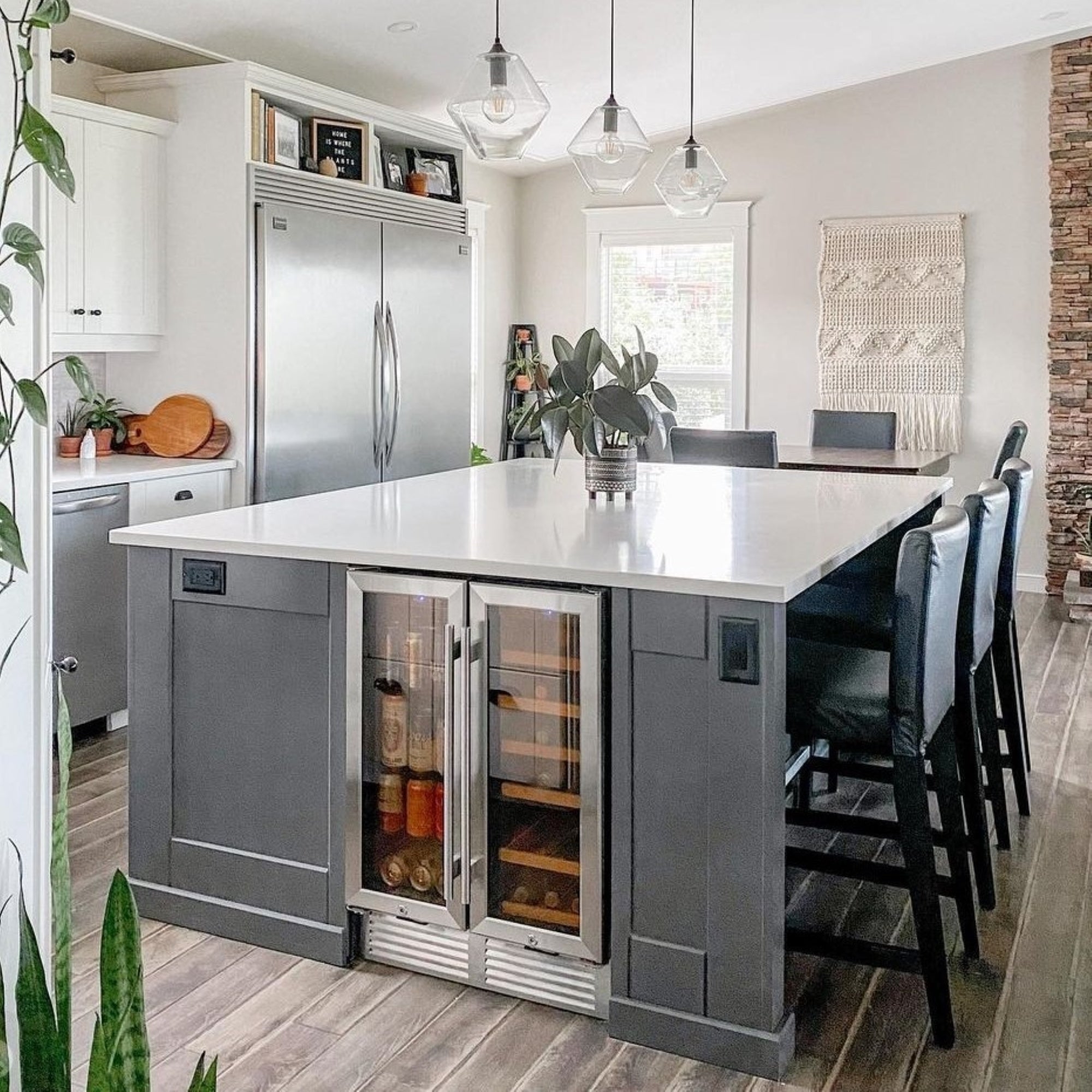 Koolatron dual zone front-venting compressor wine and beer fridge installed in a dark gray kitchen island with a white countertop and four black bar height chairs. There are pendant lamps above and white cupboards and stainless steel appliances in the background