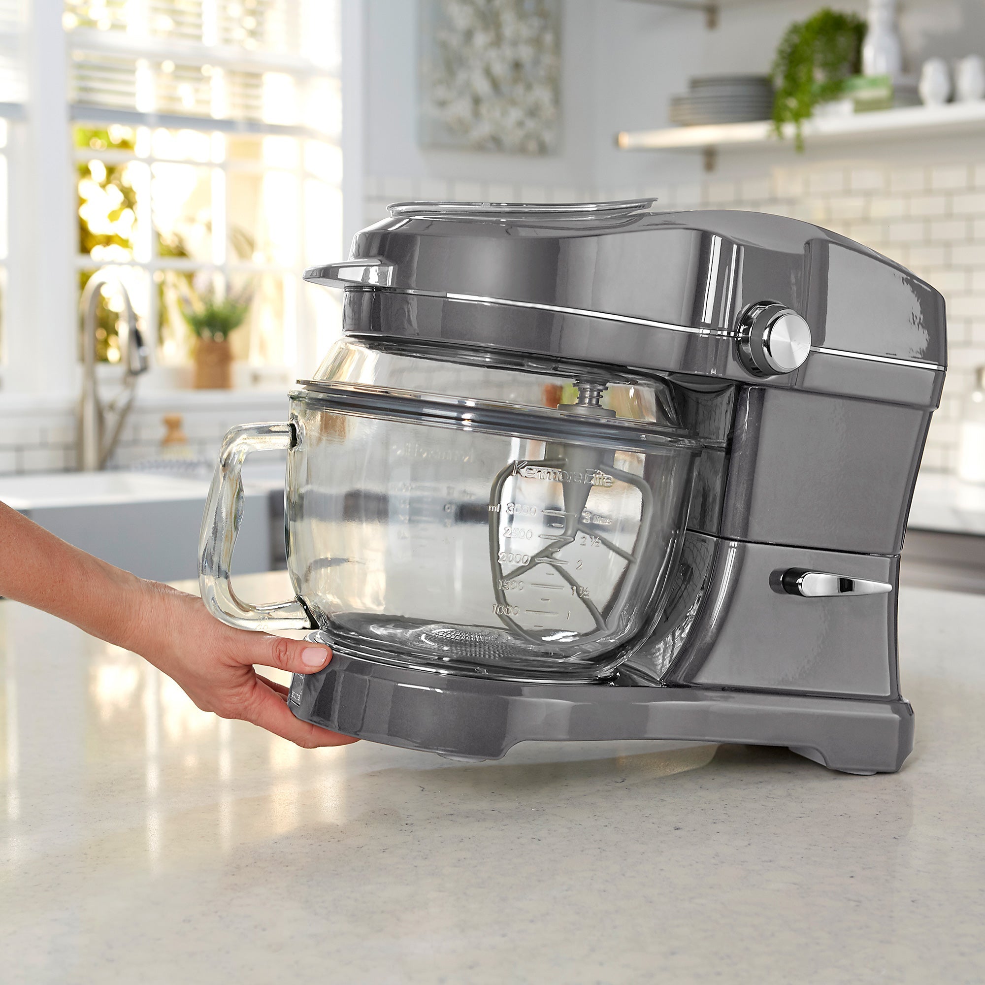 A person's hand tilting the Kenmore Elite Ovation stand mixer to move it using the tilt-and-glide feature across a light-colored kitchen counter with cupboards in the background