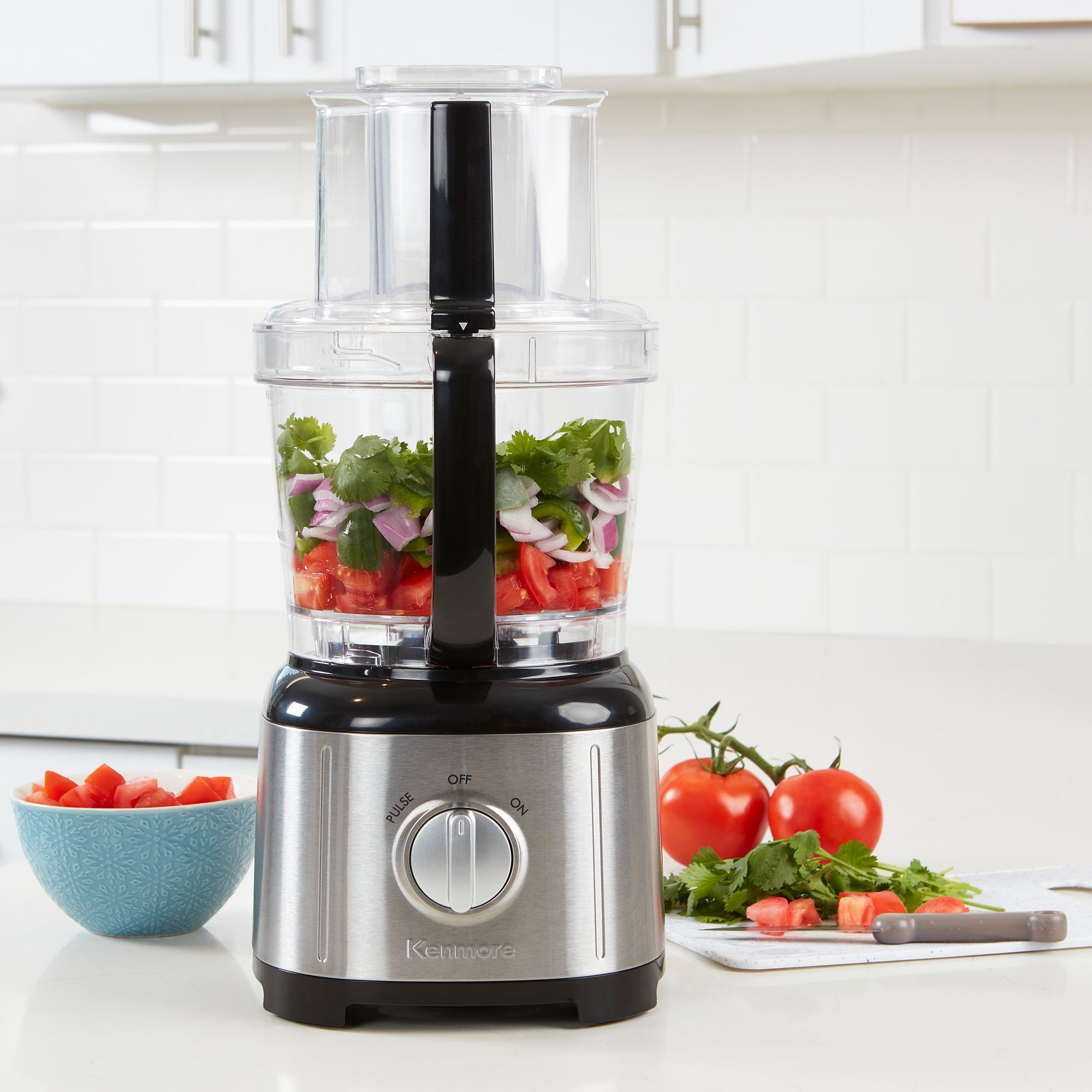Kenmore 11 cup food processor and vegetable chopper on a light gray countertop with white tile backsplash behind. There are chopped tomatoes, red onions, and cilantro in the food processor, a bowl of chopped tomatoes to the left and a cutting board, knife, and whole and chopped ingredients to the right