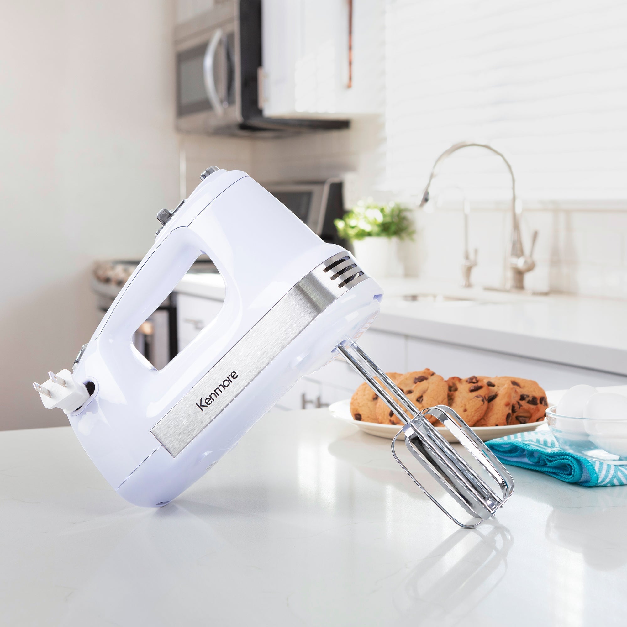 Kenmore hand mixer with beaters standing on a light gray countertop with a plate of chocolate chip cookies, a turquoise dish towel, and a glass bowl of eggs beside it and a sink, oven, and microwave in the background