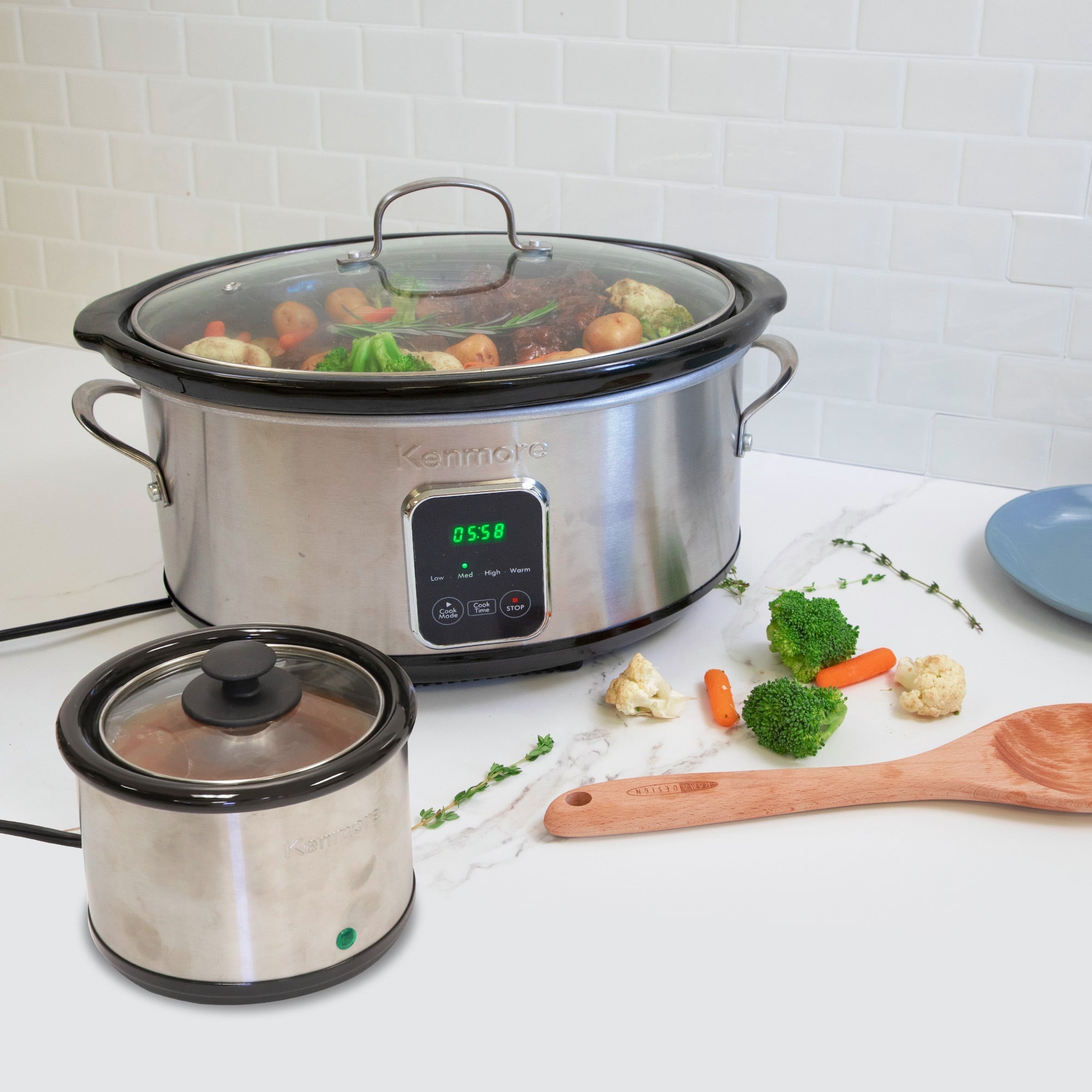 Kenmore 7 qt programmable slow cooker and sauce warmer on a white marbled countertop with white tile backsplash behind. The slow cooker contains a roast and vegetables and there are pieces of broccoli and carrots, fresh thyme sprigs, and a wooden spoon on the counter beside it