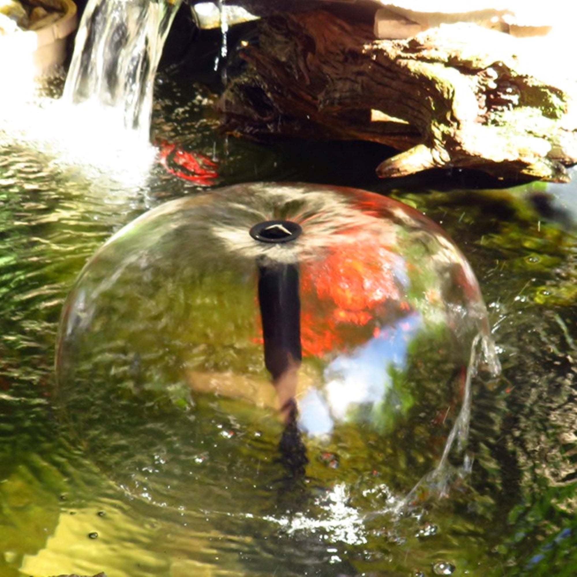 Lifestyle image of water bell fountain on the surface of a pond with a small waterfall in the background