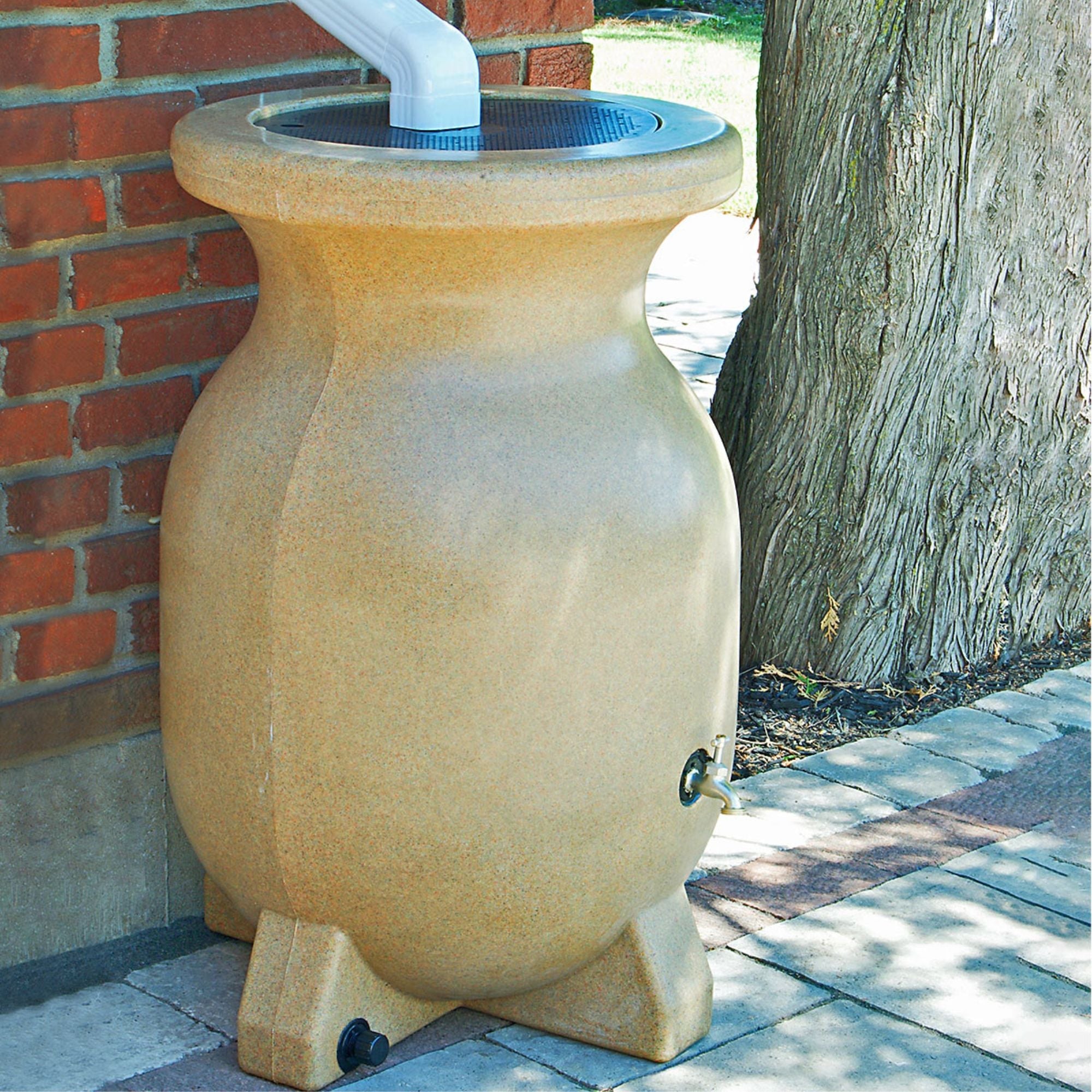Lifestyle image of rain barrel set up on a brick pathway under a white downspout with a red brick wall, a tree, and grass in the background