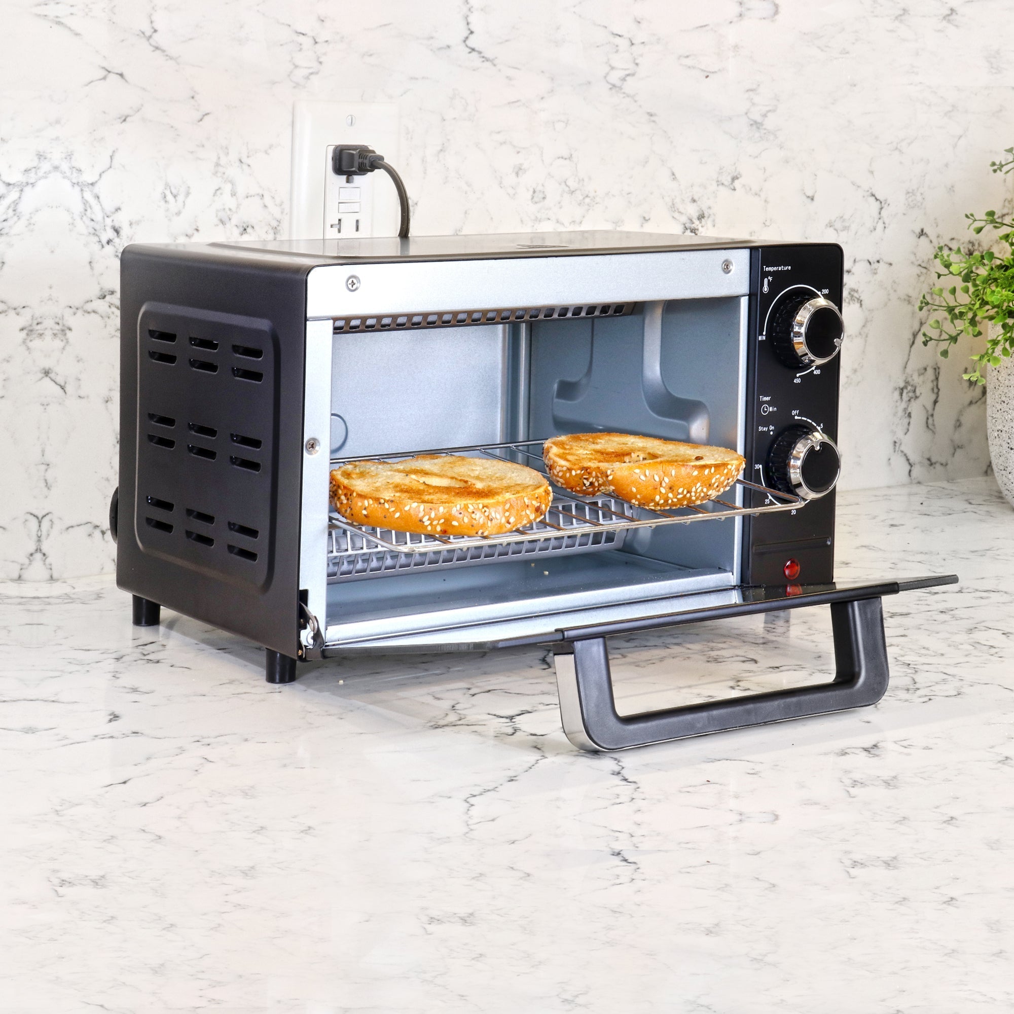 Lifestyle image of toaster oven with door open and two toasted bagel halves inside on a white marble counter with a white marble backsplash