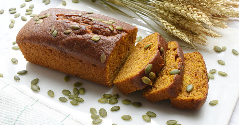 Photo of a partly sliced loaf of spiced pumpkin bread on a white cutting board with toasted pumpkin seeds sprinkled on top and around it