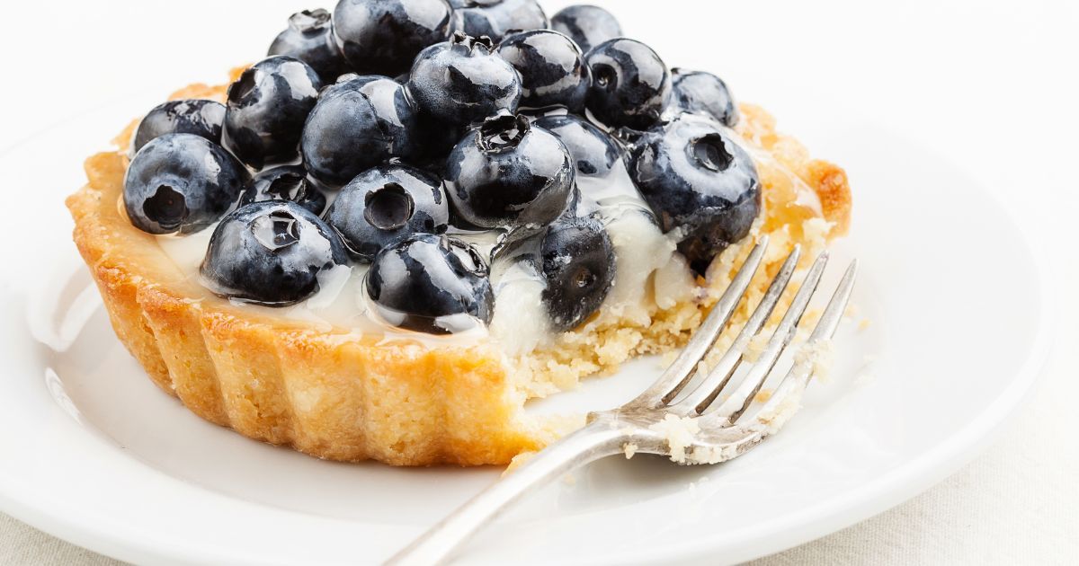 Picture of a golden brown tart shell piled with creamy filling and fresh blueberries on a white plate with a fork