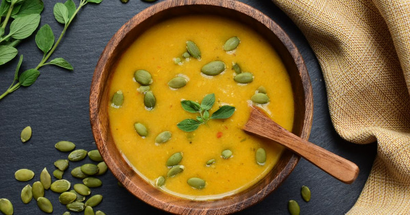Photo of a wooden bowl filled with thick and creamy orange colored soup with pumpkin seeds and fresh herbs sprinkled on top and around it