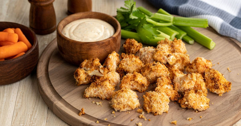 Photo of golden brown breaded cauliflower florets on a wooden cutting board with a bowl of baby carrots, a bowl of dipping sauce, and a pile of celery sticks arranged around them