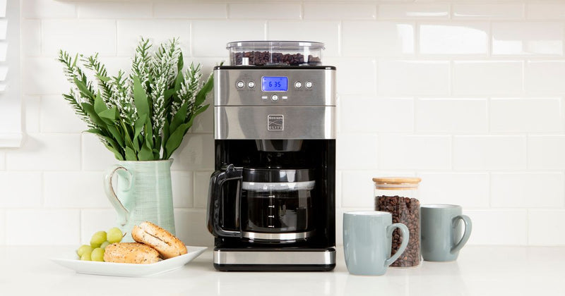 Photo of the Kenmore Grind and Brew coffee maker on a white kitchen counter with light blue coffee mugs and a plant arranged around it