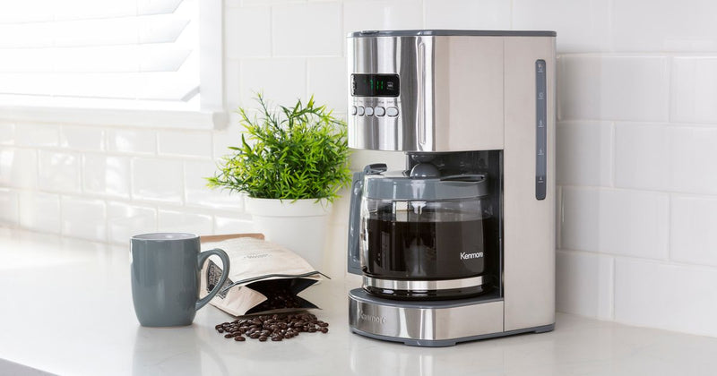 A white coffee maker with a full coffee pot sits on a white countertop. Next to the coffee maker is a green plant in a white pot, a bag of coffee beans spilled over revealing the beans, and a white coffee mug.