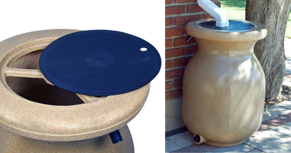 A stone looking rain barrel sits next to the brick wall of a home. The downspout is located directly above the rain barrel allowing the water to pour into it. Next to the rain barrel is the trunk of a tree.