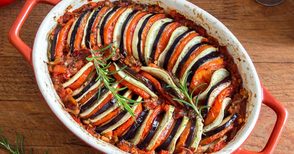 Picture of an oval ceramic dish filled with thinly sliced vegetables (tomotoes, potatoes, zucchini) arranged in an alternating pattern with fresh herbs on top
