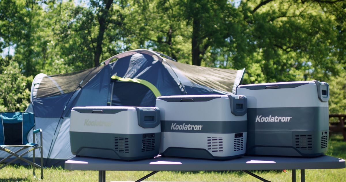 Picture of a campsite with Koolatron's SK30 30 liter capacity, SK40 40 liter capacity, and SK50 50 liter capacity coolers on a folding table in the foreground and a dome tent, folding camping chair, and trees in the background