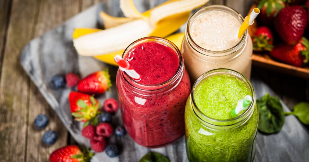 Photo of three mason jars containing a purple-red berry smoothie, a cream-coloured almond-peach smoothie, and a bright green smoothie with matching straws