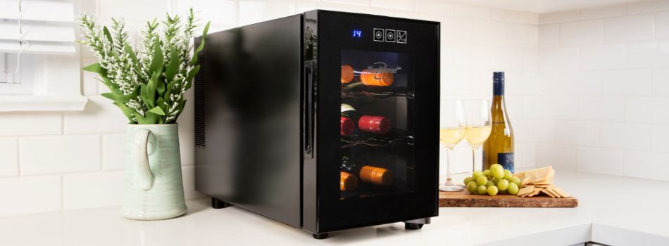 Lifestyle image of 6 bottle wine fridge on a white counter with a white tile backsplash behind, a pitcher of flowers to the left, and 2 wine glasses, a wine bottle, grapes and cheese set on a wooden chopping board on the right