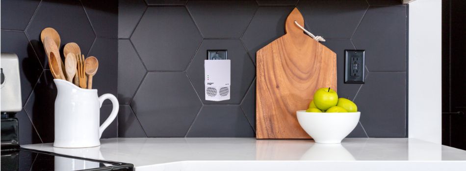 Photo of a contemporary-style white kitchen counter with dark gray tile backsplash, bowl of fruit, wooden cutting board, and a white rodent repeller plugged in