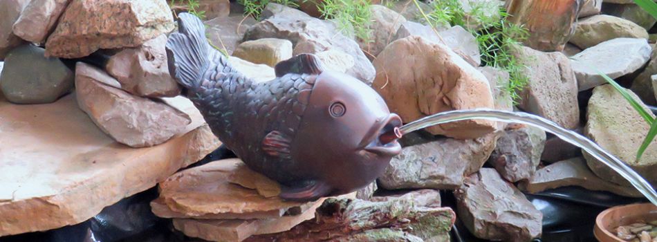 Lifestyle image of bronze-look fish spitter set up on a flat rock at the side of a pond