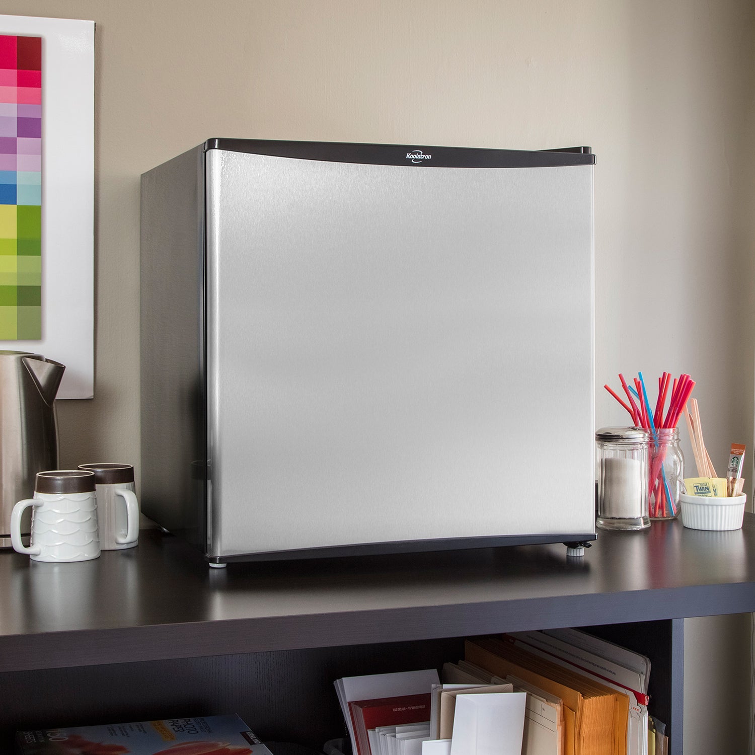 Black and stainless steel compact fridge with freezer on a dark gray countertop of a coffee bar with a kettle and mugs to the left and sugar and stir sticks to the right.