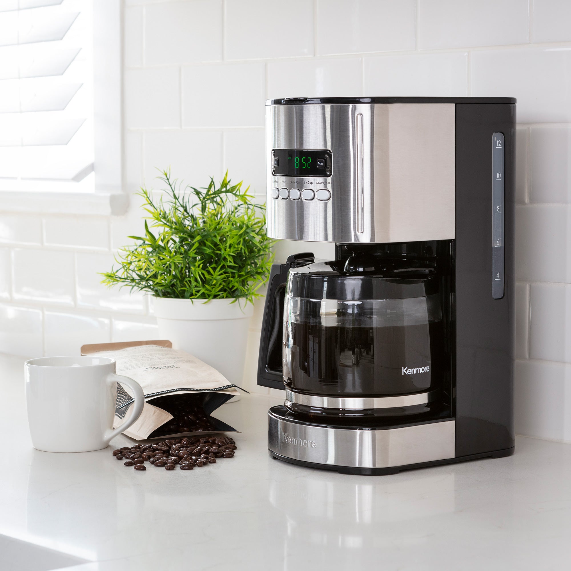 Kenmore 12 cup programmable coffeemaker on a light gray countertop with white tile backsplash behind and a mug, bag of coffee beans, and potted plant beside it