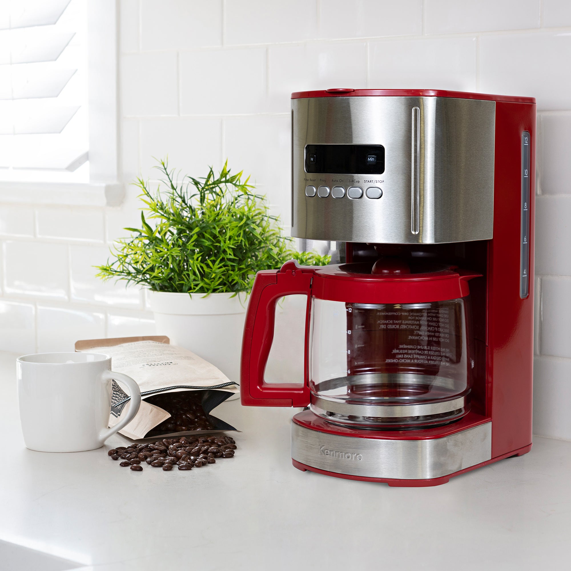 Kenmore 12 cup programmable coffeemaker on a light gray countertop with white tile backsplash behind and a mug, bag of coffee beans, and potted plant beside it