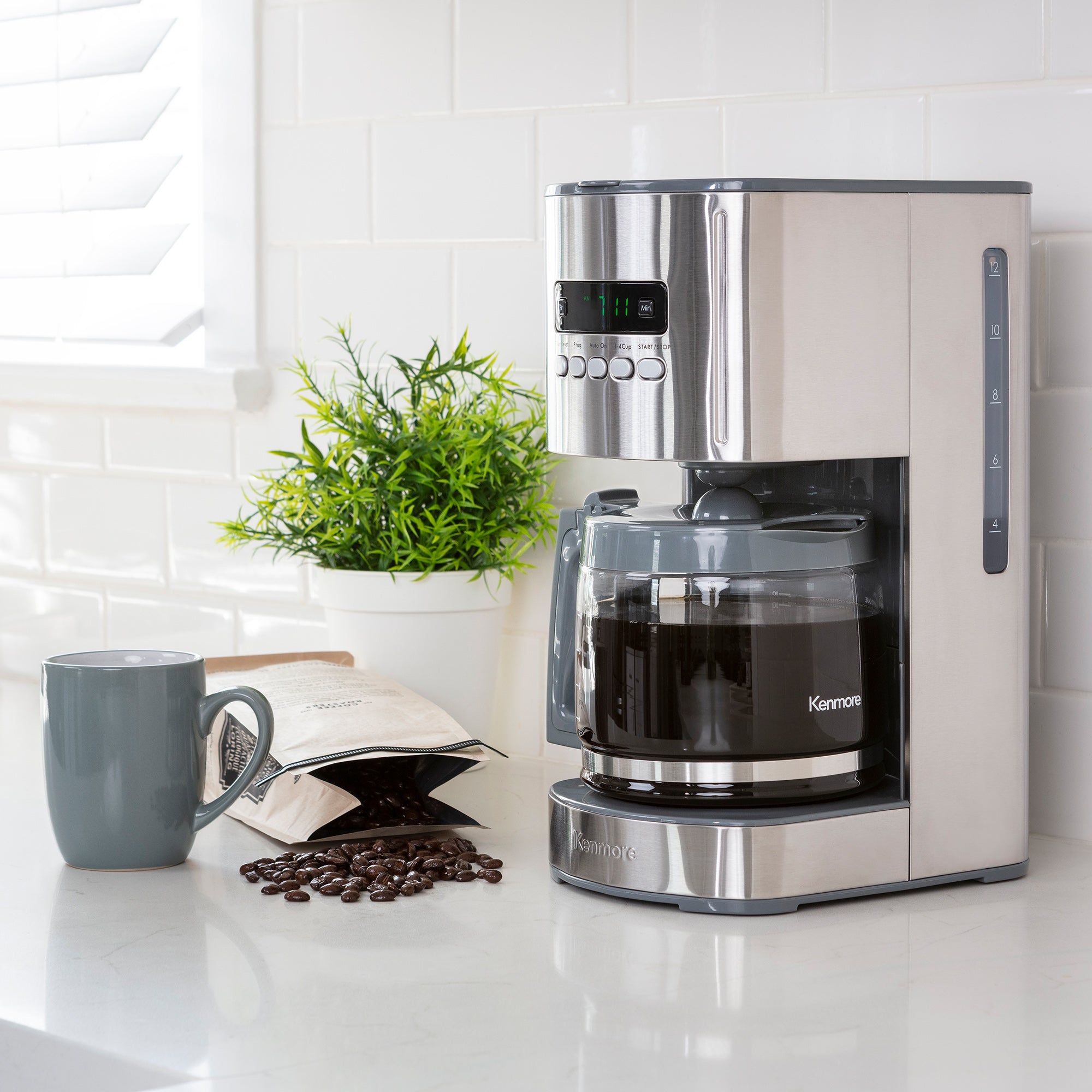 Kenmore 12 cup programmable coffeemaker on a light gray countertop with white tile backsplash behind and a mug, bag of coffee beans, and potted plant beside it