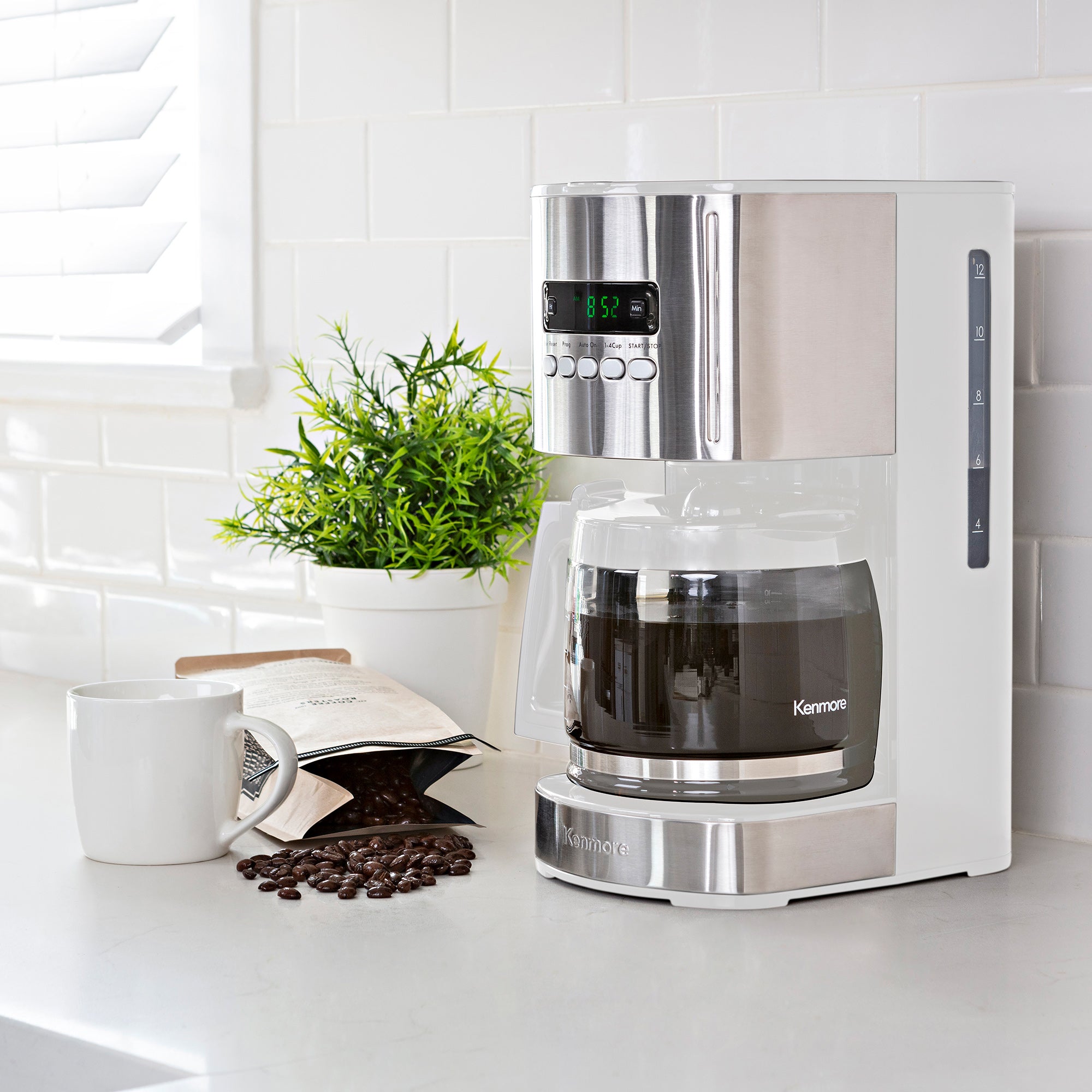 Kenmore 12 cup programmable coffeemaker on a light gray countertop with white tile backsplash behind and a mug, bag of coffee beans, and potted plant beside it