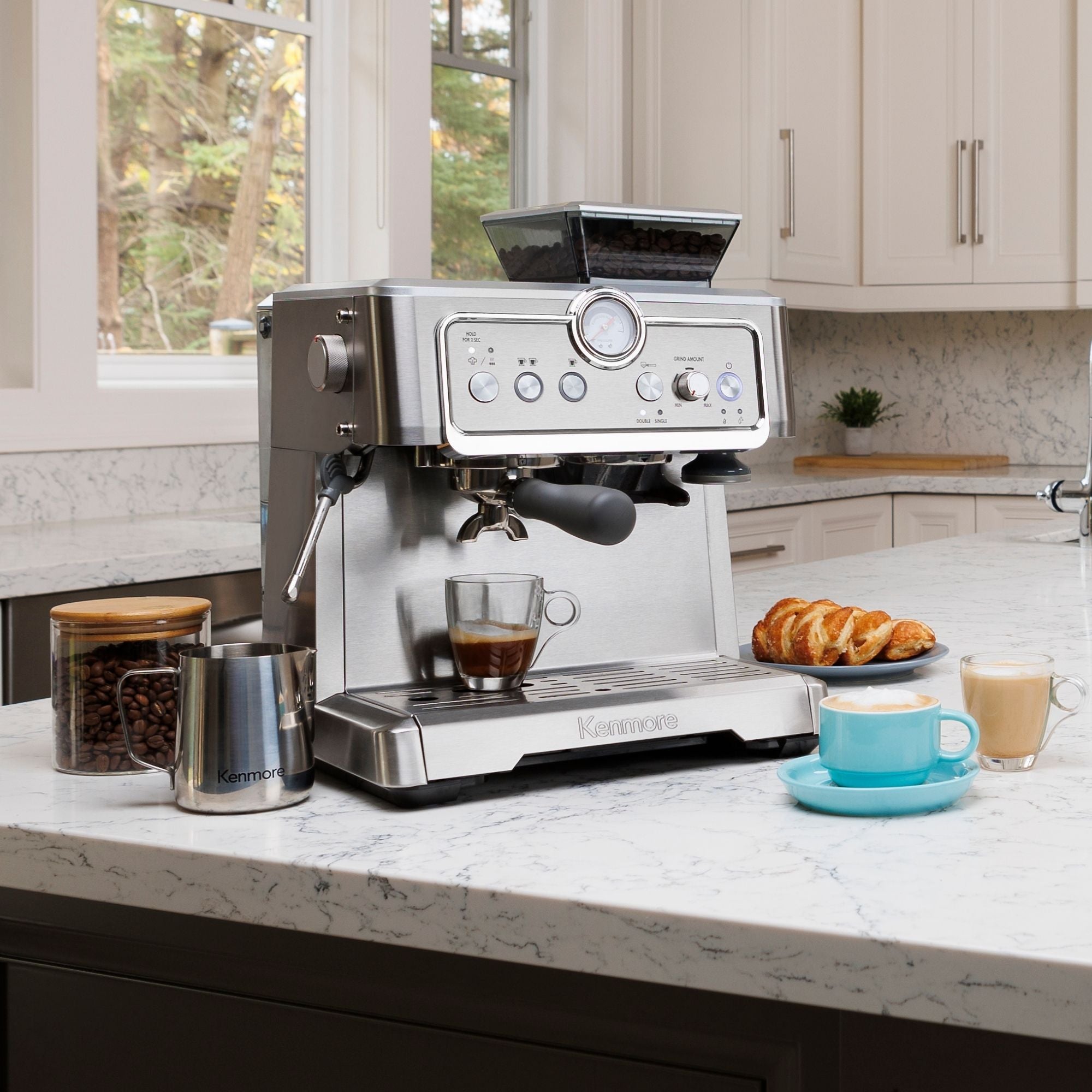 Kenmore semi-automatic espresso maker with espresso being extracted into two clear cups on a white marble countertop in a bright white kitchen. A jar of whole coffee beans, stainless steel milk pitcher, a pastry, a latte in a clear mug, and a cappuccino in a light blue mug are arranged around it.