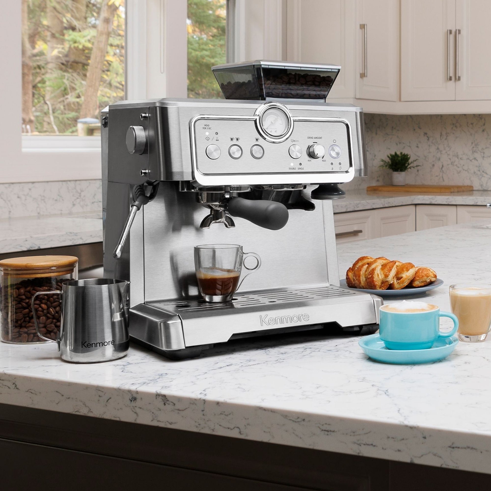 Kenmore semi-automatic espresso maker with espresso being extracted into two clear cups on a white marble countertop in a bright white kitchen. A jar of whole coffee beans, stainless steel milk pitcher, a pastry, a latte in a clear mug, and a cappuccino in a light blue mug are arranged around it.