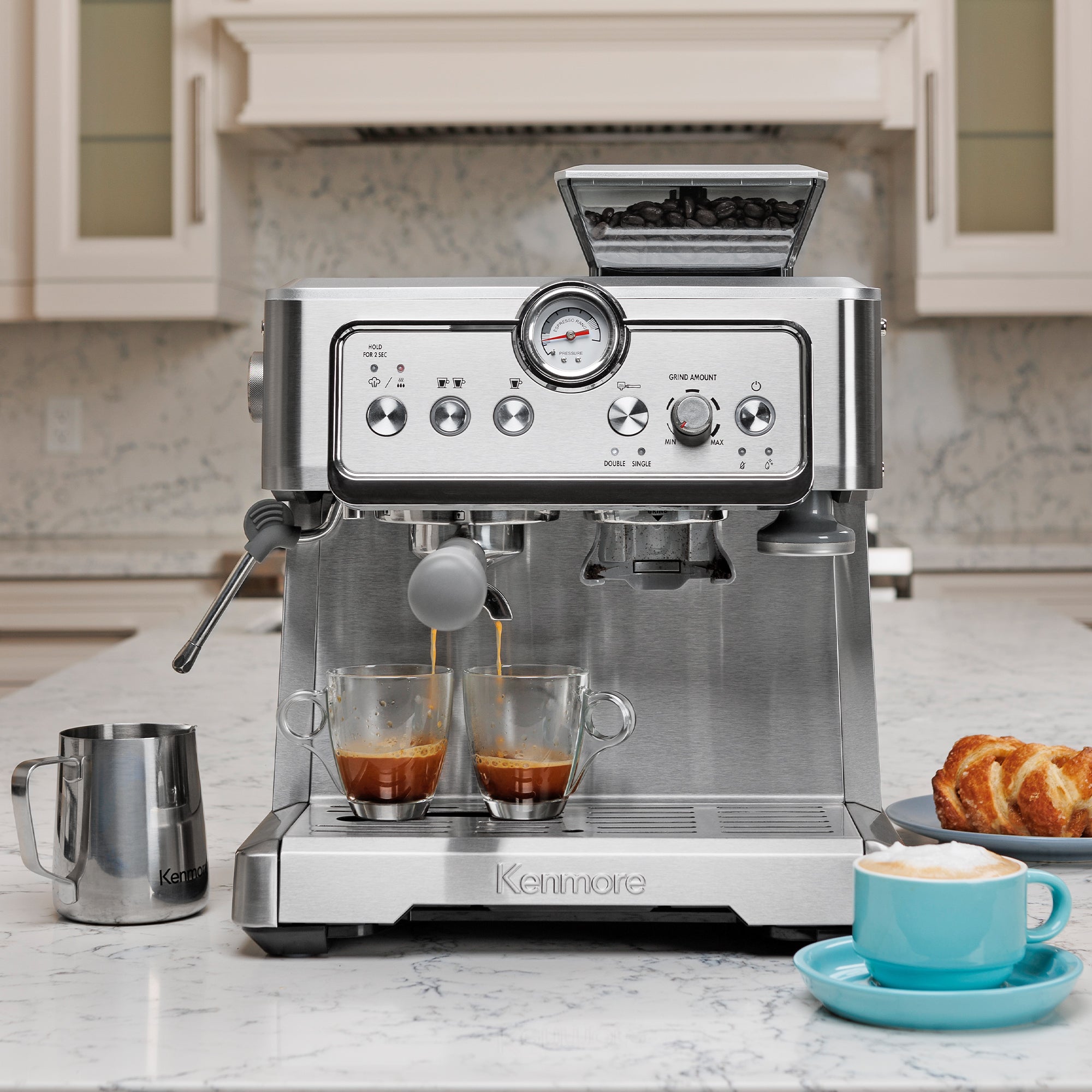 Kenmore semi-automatic espresso maker with espresso being extracted into two clear cups on a white marble countertop with the milk pitcher, a pastry, and a cappuccino in a light blue mug arranged around it.