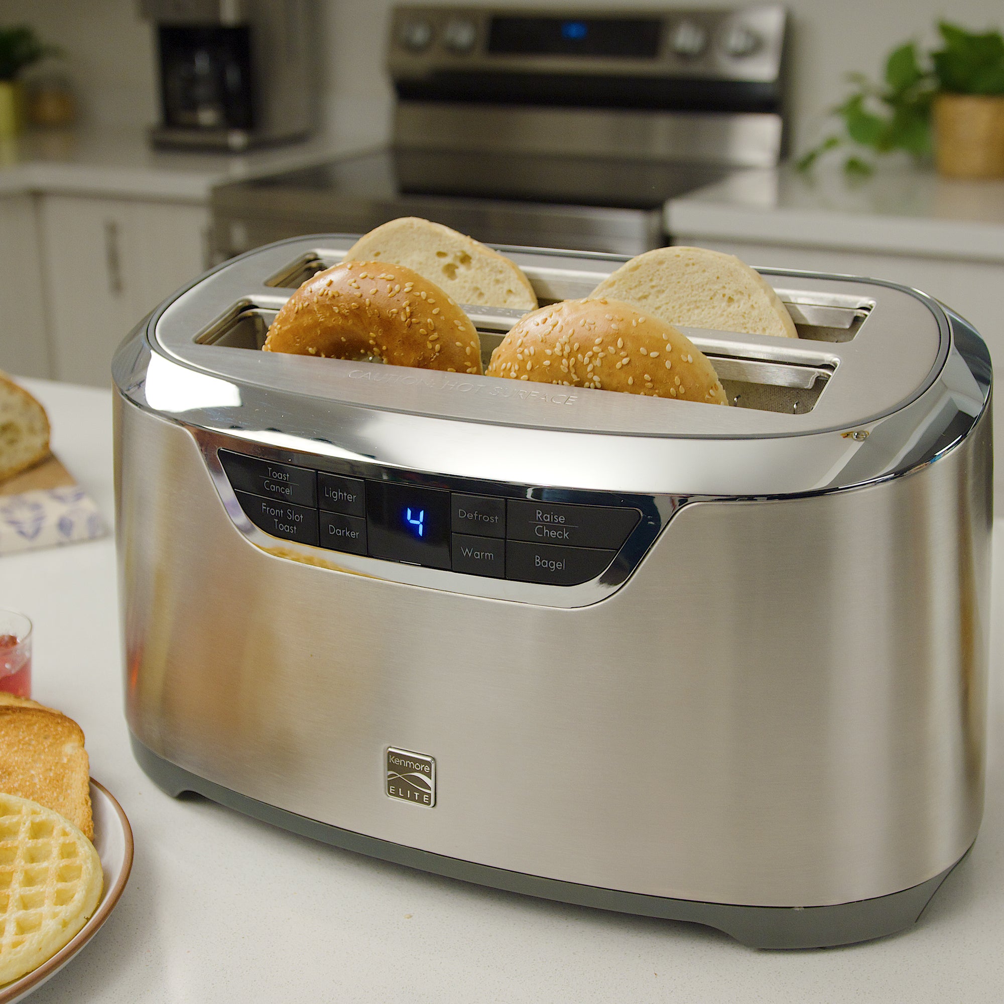 Kenmore Elite long slot stainless steel toaster with four toasted bagel halves inside on a light gray countertop with a stainless steel stove and potted plant in the background
