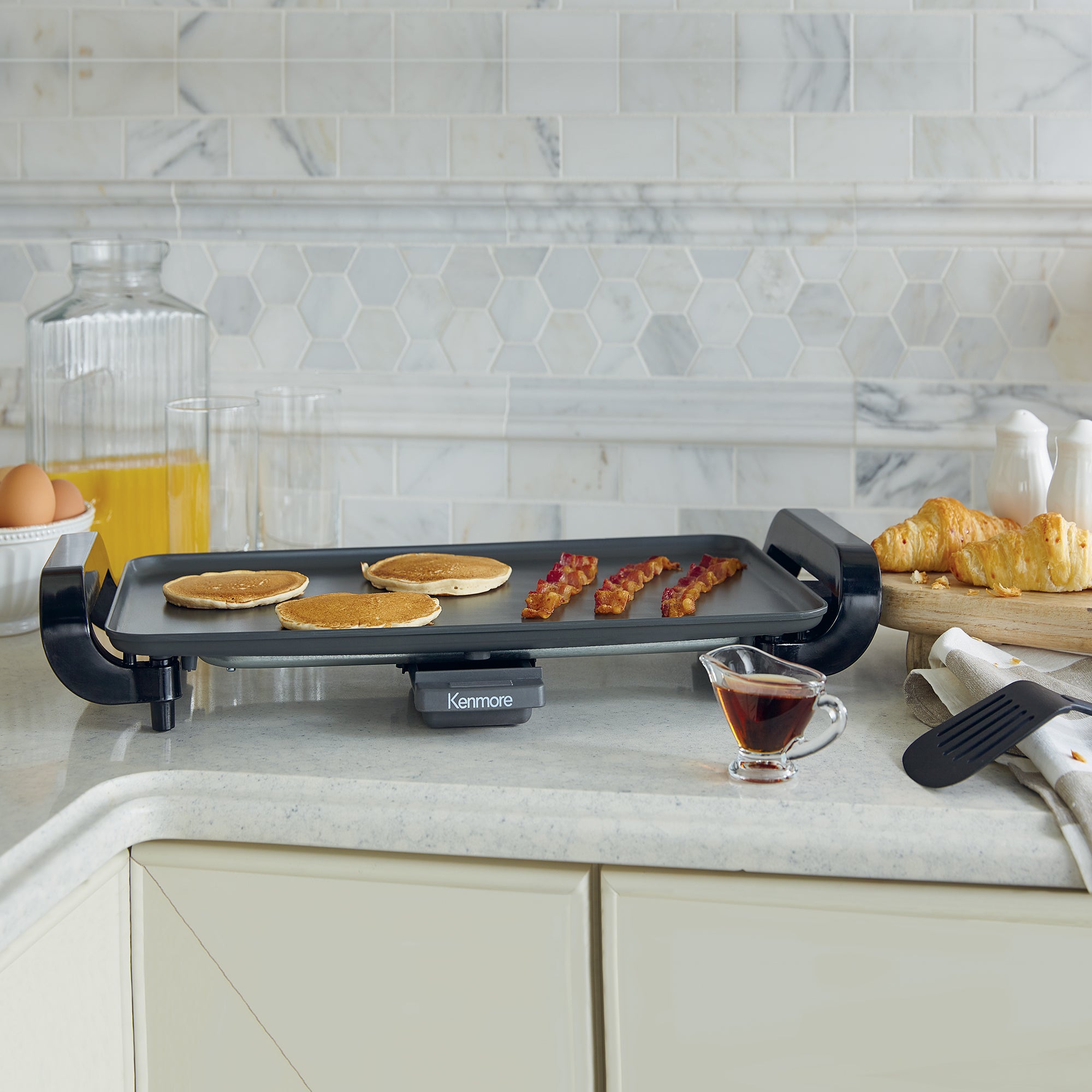 Kenmore non-stick electric griddle with pancakes and bacon cooking on a white countertop with light gray tile backsplash behind and orange juice, brown eggs, croissants, a small glass container of syrup, and a spatula arranged around it