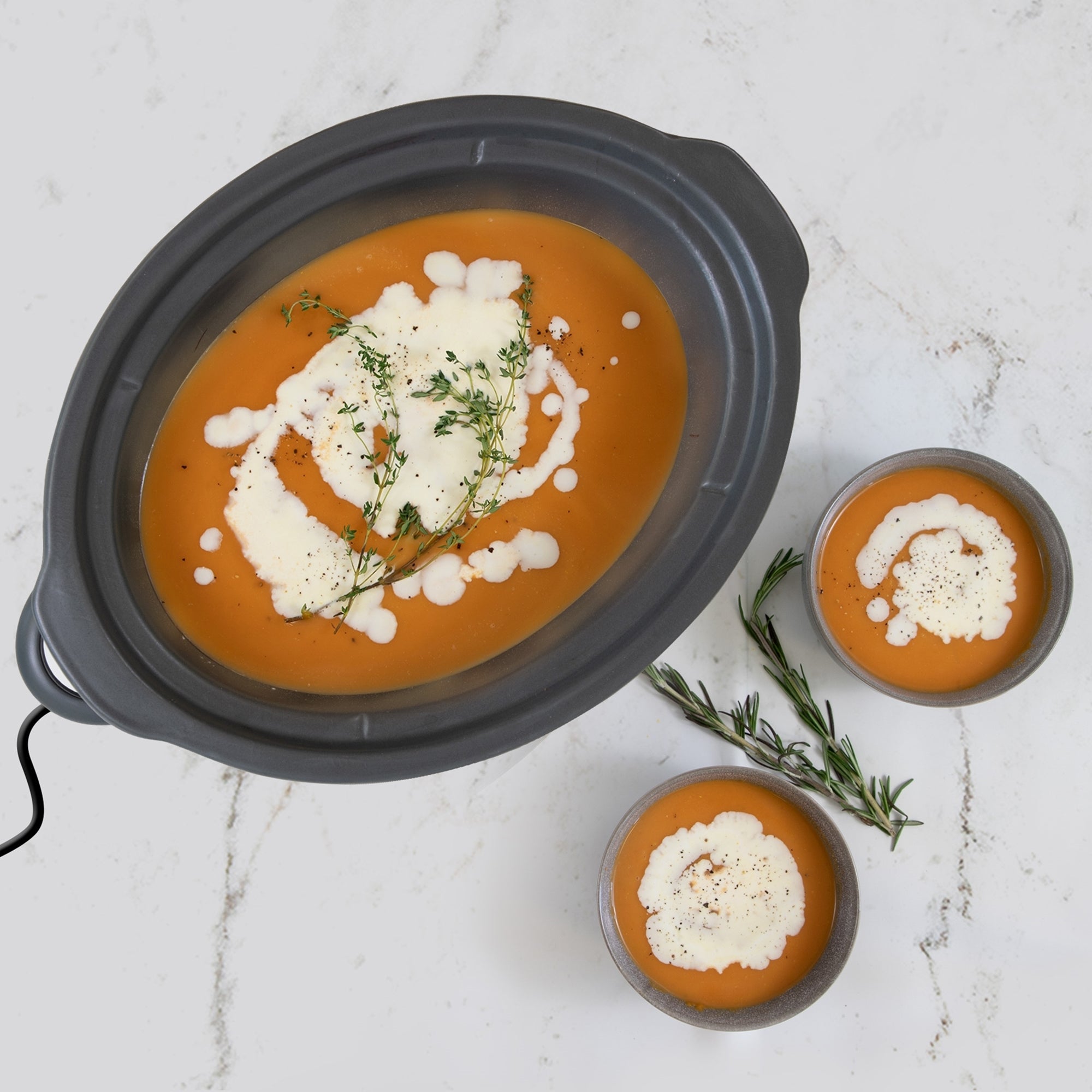 Kenmore 5 quart black slow cooker without lid viewed from above on a white marbled countertop. The slow cooker is filed with orange colored soup with cream drizzled on top and there are two bowls of soup and sprigs of fresh thyme beside it