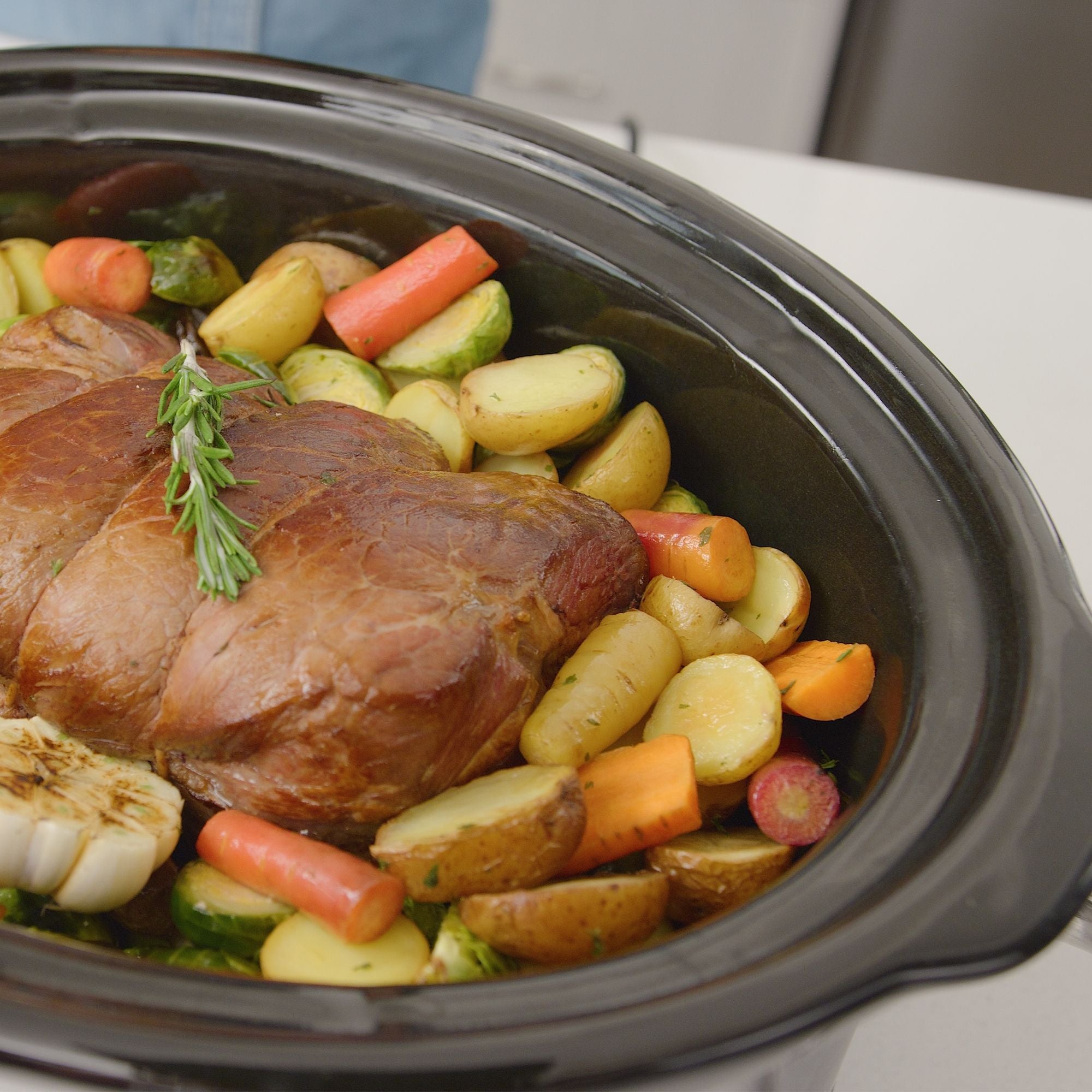 Closeup of a beef roast on a bed of carrots, potatoes, and brussels sprouts with roasted garlic and a sprig of fresh rosemary in the 7-qt Kenmore programmable slow cooker