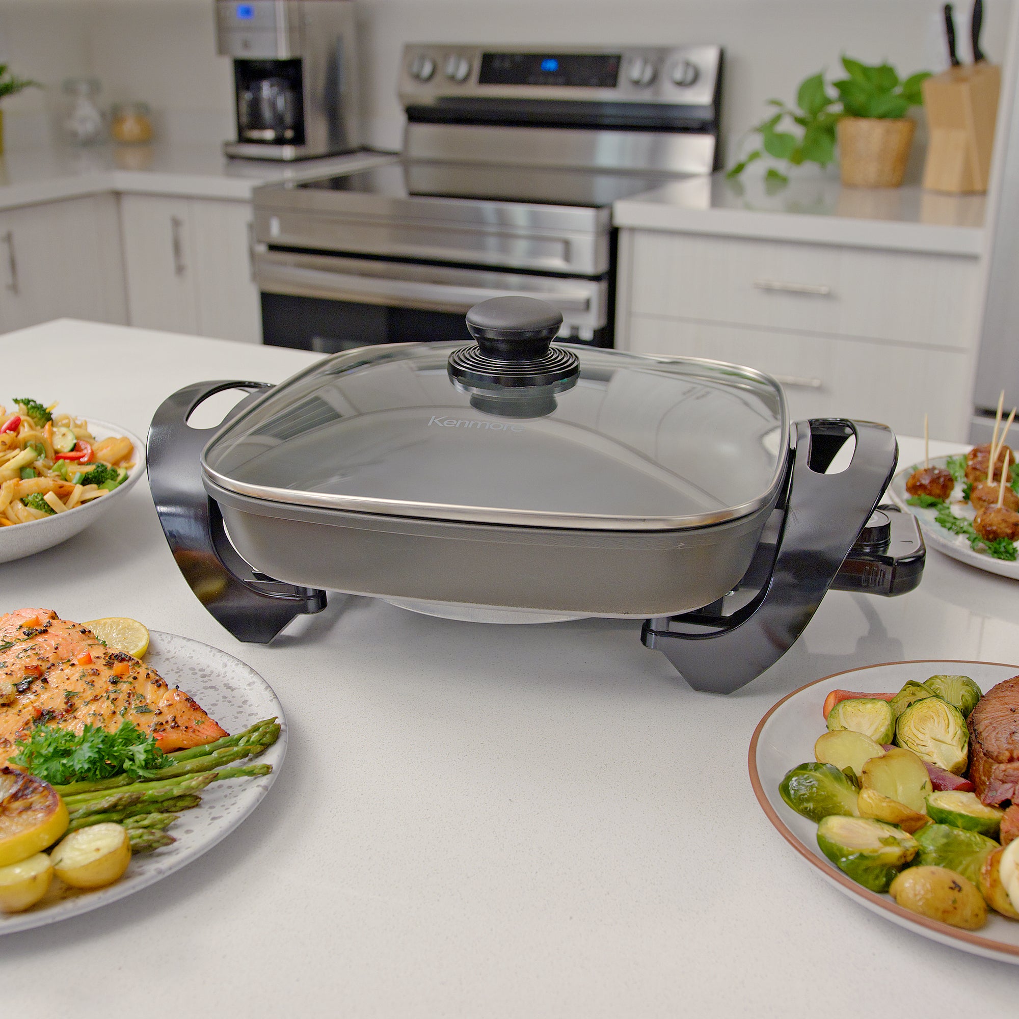 Kenmore non-stick electric skillet with glass lid on white marble kitchen island surrounded by dishes of food