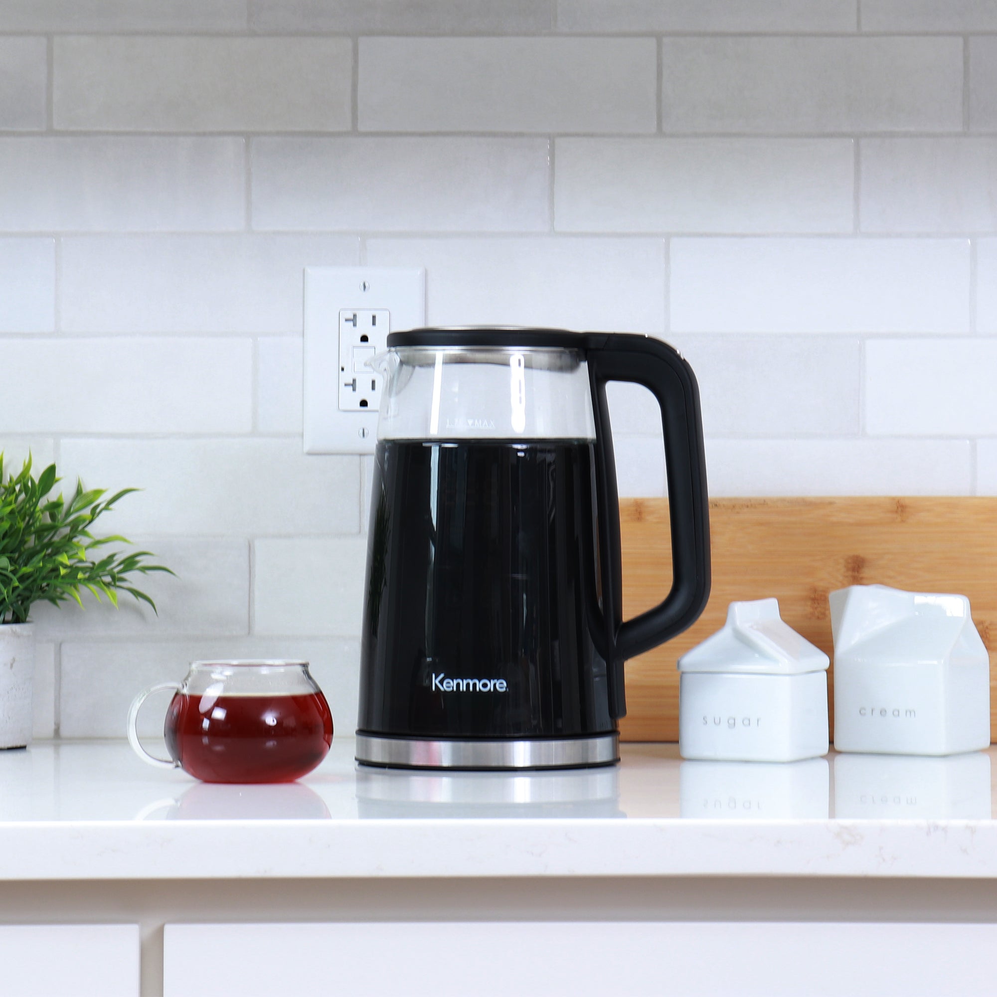 Kenmore cool-touch glass tea kettle with 4 pre-set temperatures on a white counter with a cup of tea beside it and a white tile backsplash behind