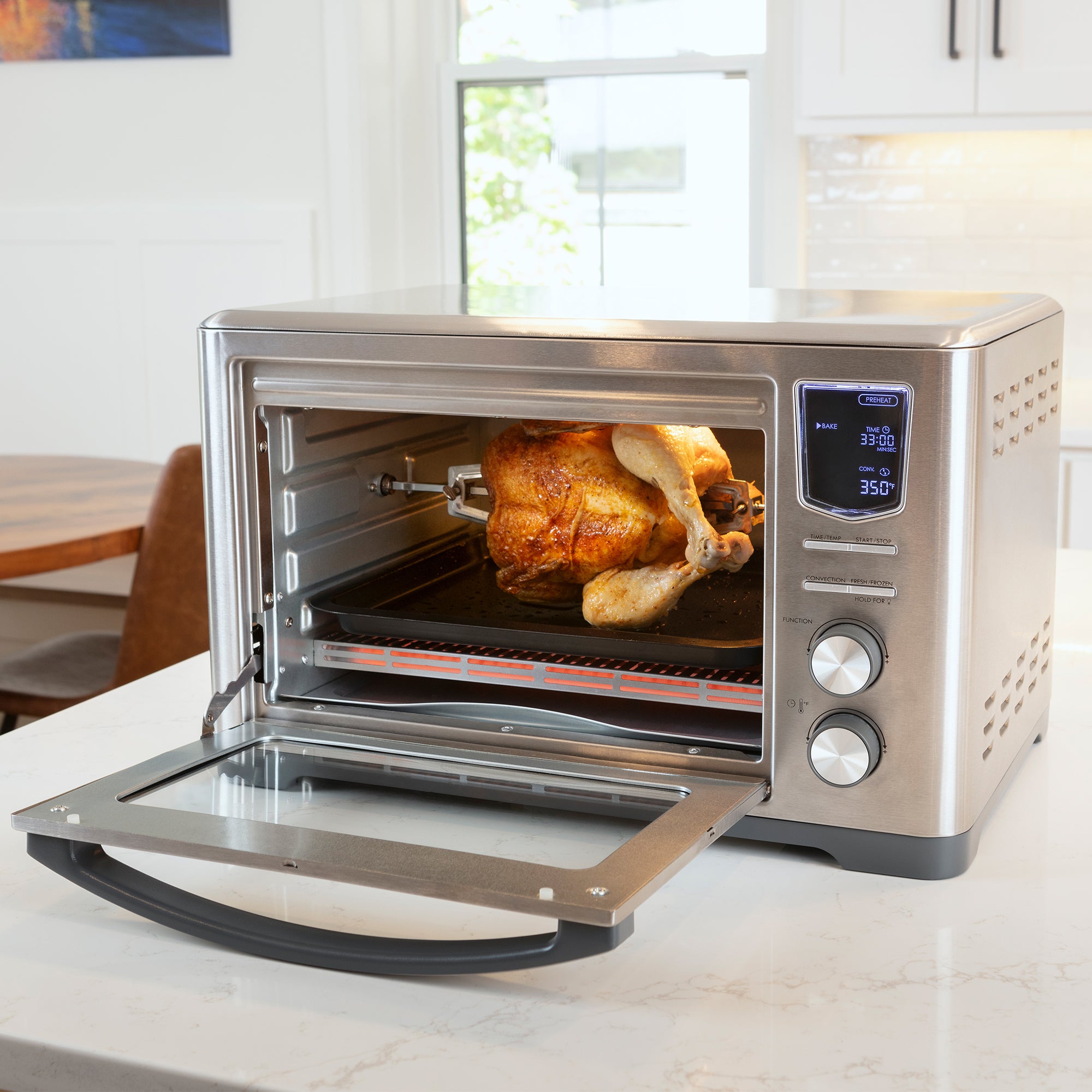 Kenmore 24 liter 11-in-1 toaster oven/air fryer/dehydrator with convection fan, open with a whole chicken on the rotisserie, on a white marble kitchen counter
