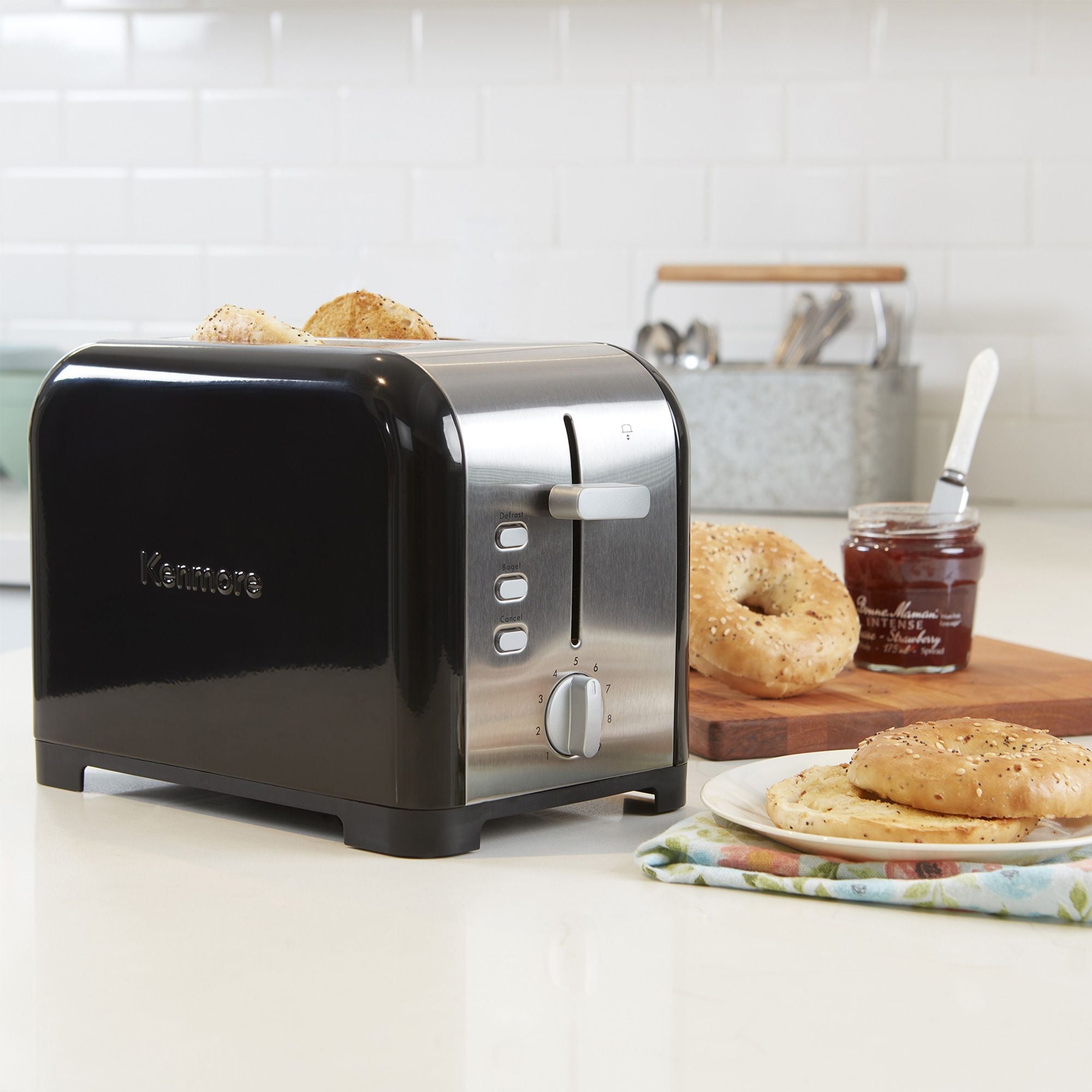 Kenmore 2-slice stainless steel toaster with toasted bagel slices inside on a light gray countertop with white tile backsplash behind. To the right of the toaster is a napkin and plate with toasted bagel and a wooden cutting board with uncut bagels and an open jam jar