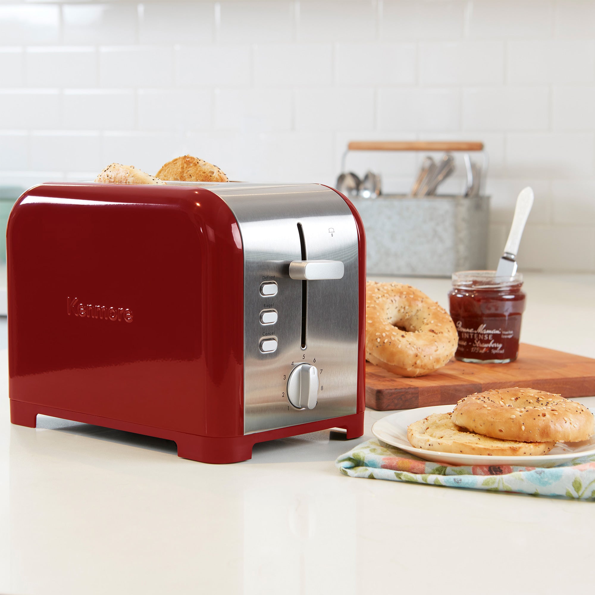 Kenmore 2-slice stainless steel toaster with toasted bagel slices inside on a light gray countertop with white tile backsplash behind. To the right of the toaster is a napkin and plate with toasted bagel and a wooden cutting board with uncut bagels and an open jam jar