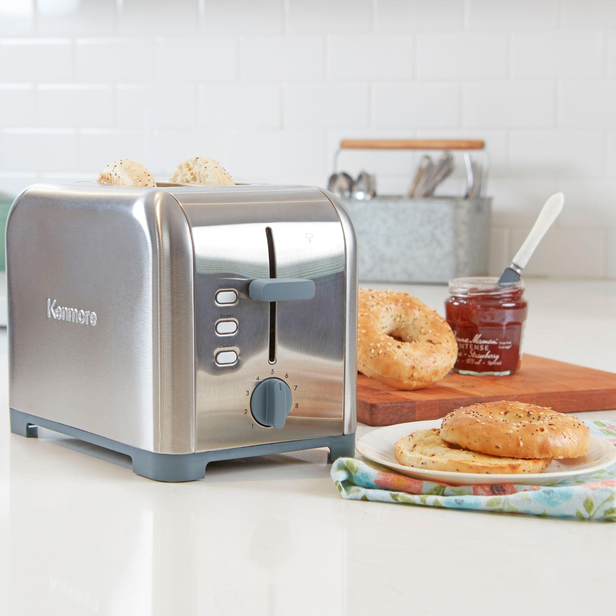 Kenmore 2-slice stainless steel toaster with toasted bagel slices inside on a light gray countertop with white tile backsplash behind. To the right of the toaster is a napkin and plate with toasted bagel and a wooden cutting board with uncut bagels and an open jam jar.
