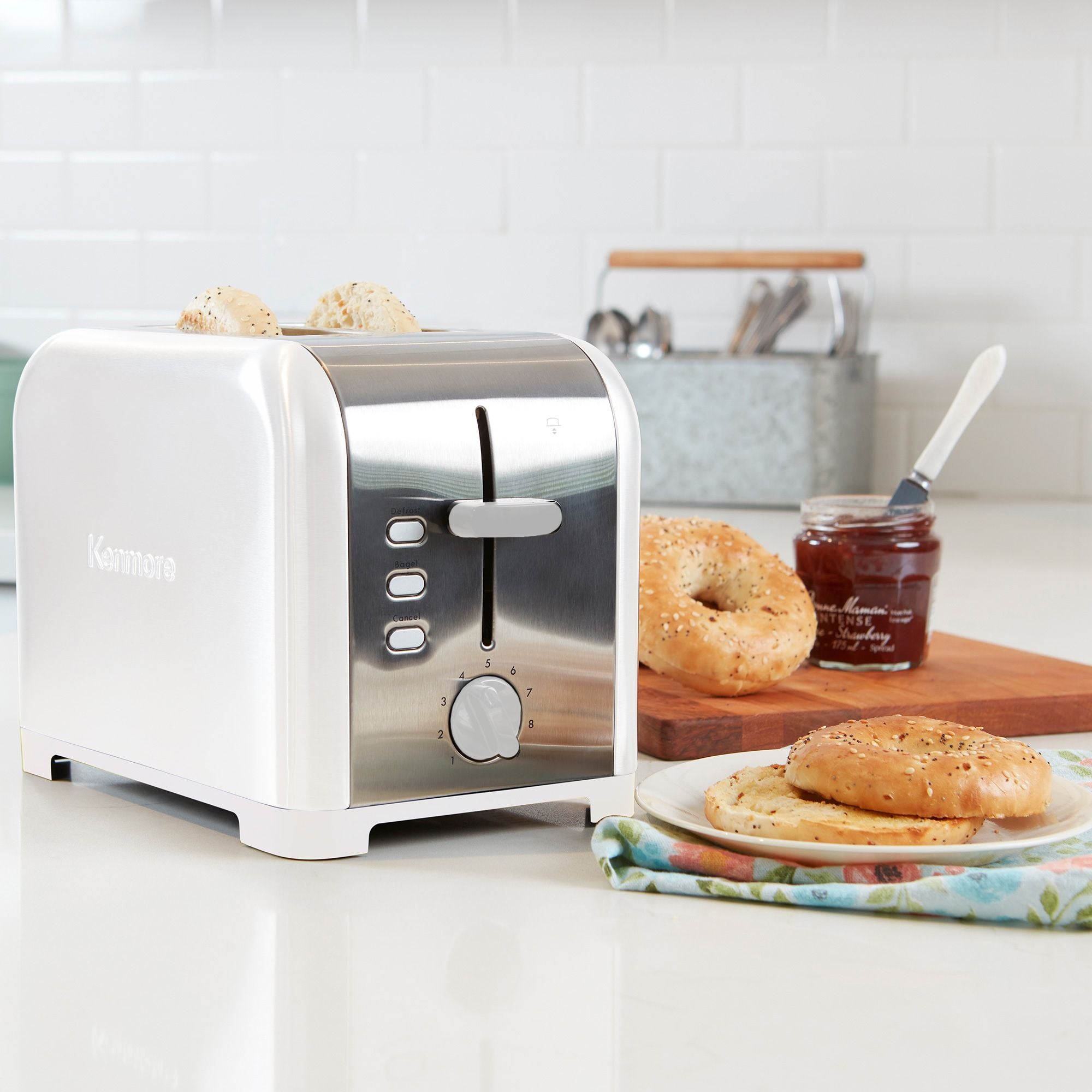 Kenmore 2-slice stainless steel toaster with toasted bagel slices inside on a light gray countertop with white tile backsplash behind. To the right of the toaster is a napkin and plate with toasted bagel and a wooden cutting board with uncut bagels and an open jam jar.