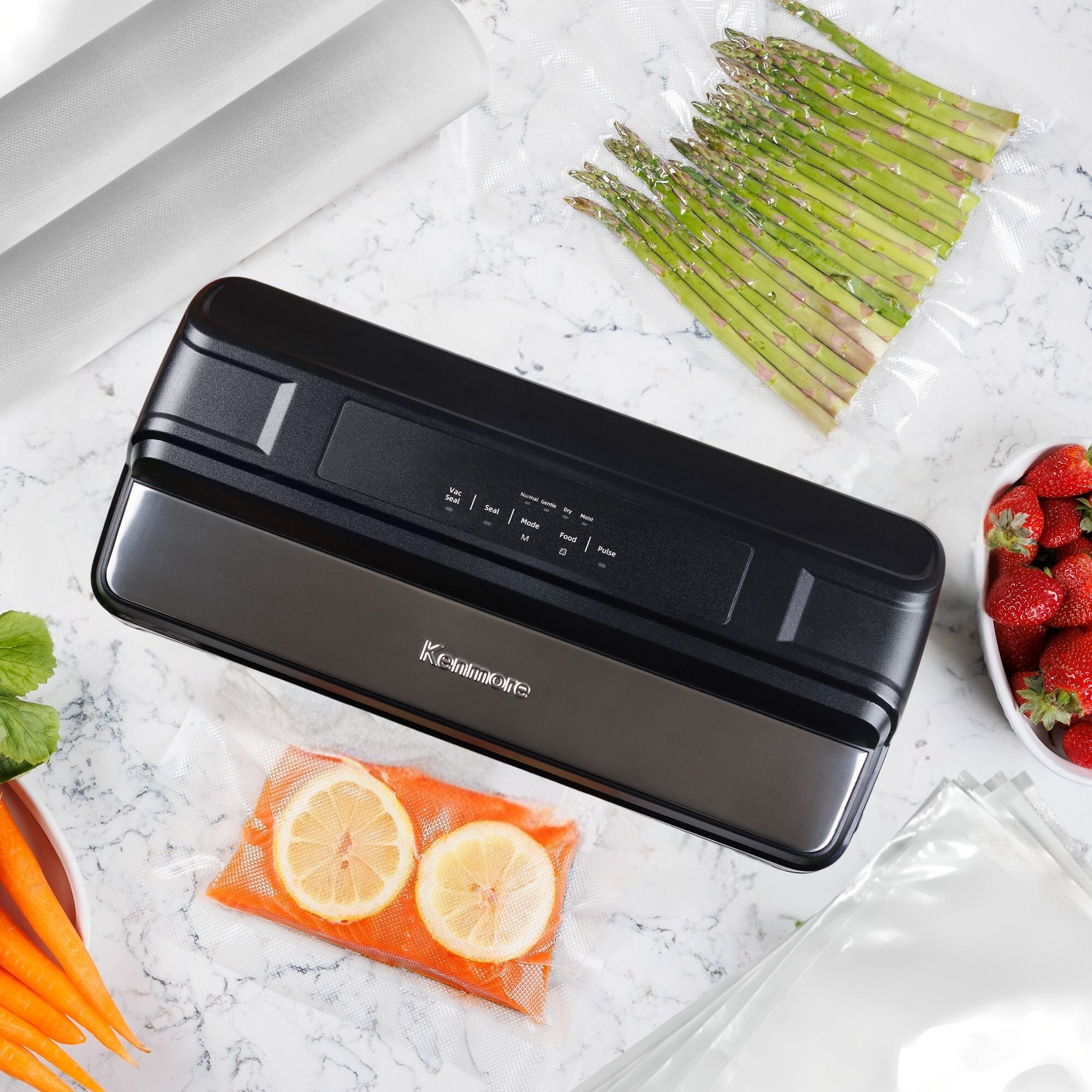 Kenmore vacuum sealer on a white and gray marble surface with a bag containing salmon and lemon slices being sealed and a sealed bag of asparagus, fresh strawberries and carrots, and vacuum bags and bag rolls arranged around it