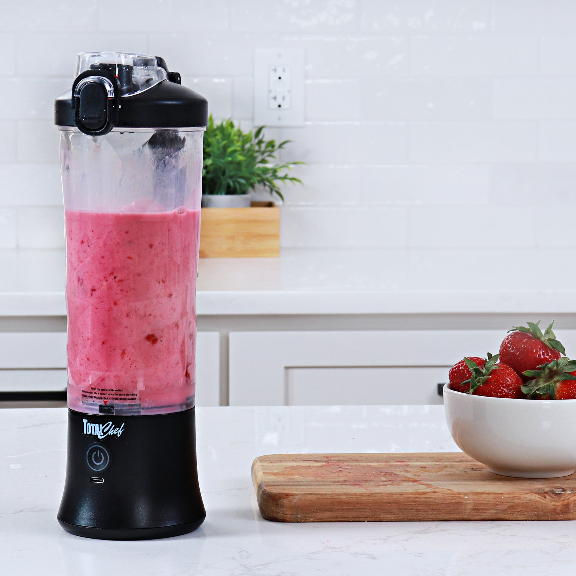Lifestyle image of the Total Chef portable rechargeable blender filled with bright pink smoothie in a white kitchen with a wooden cutting board and a white bowl of strawberries beside it.