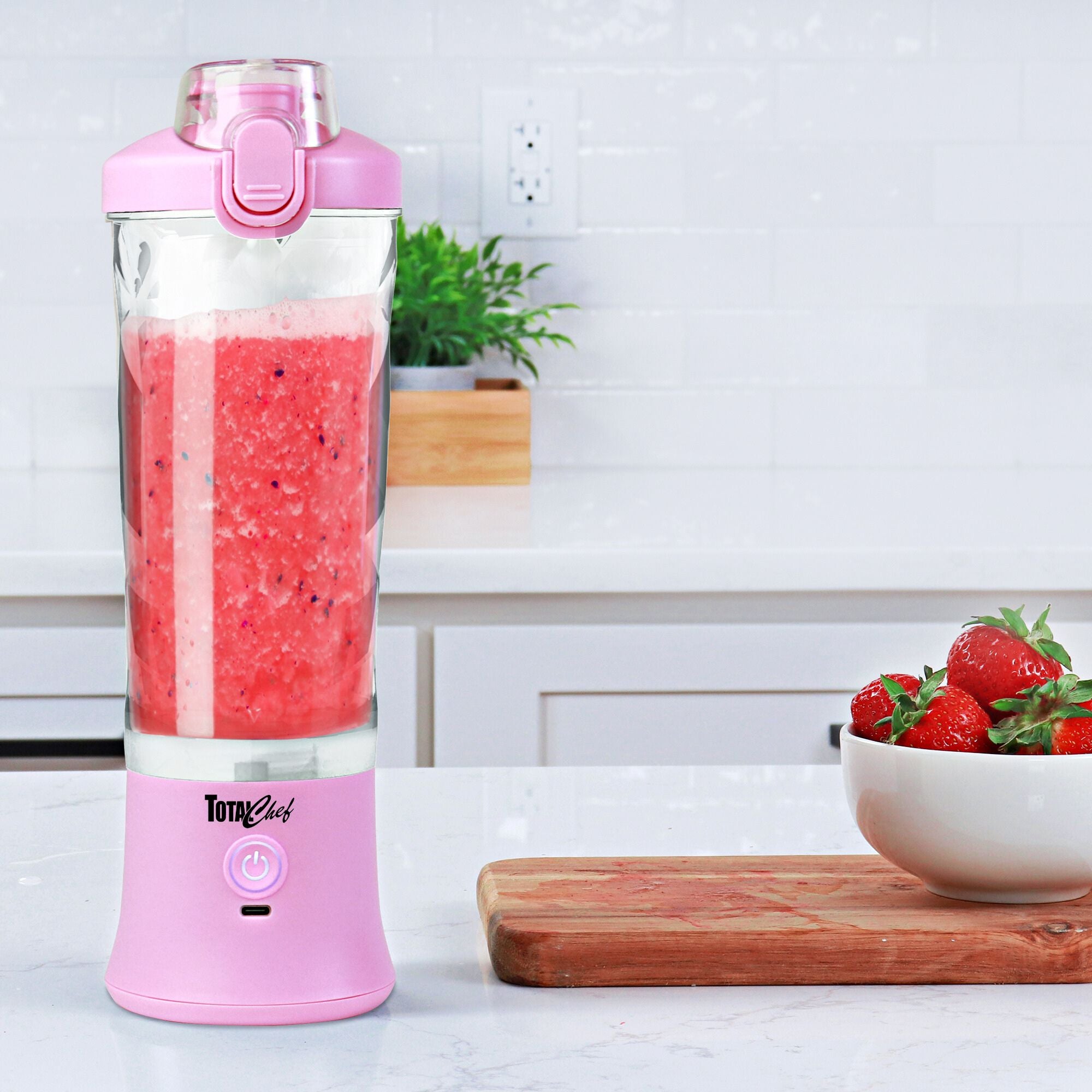 Lifestyle image of the Total Chef portable rechargeable blender filled with bright pink smoothie in a white kitchen with a wooden cutting board and a white bowl of strawberries beside it.