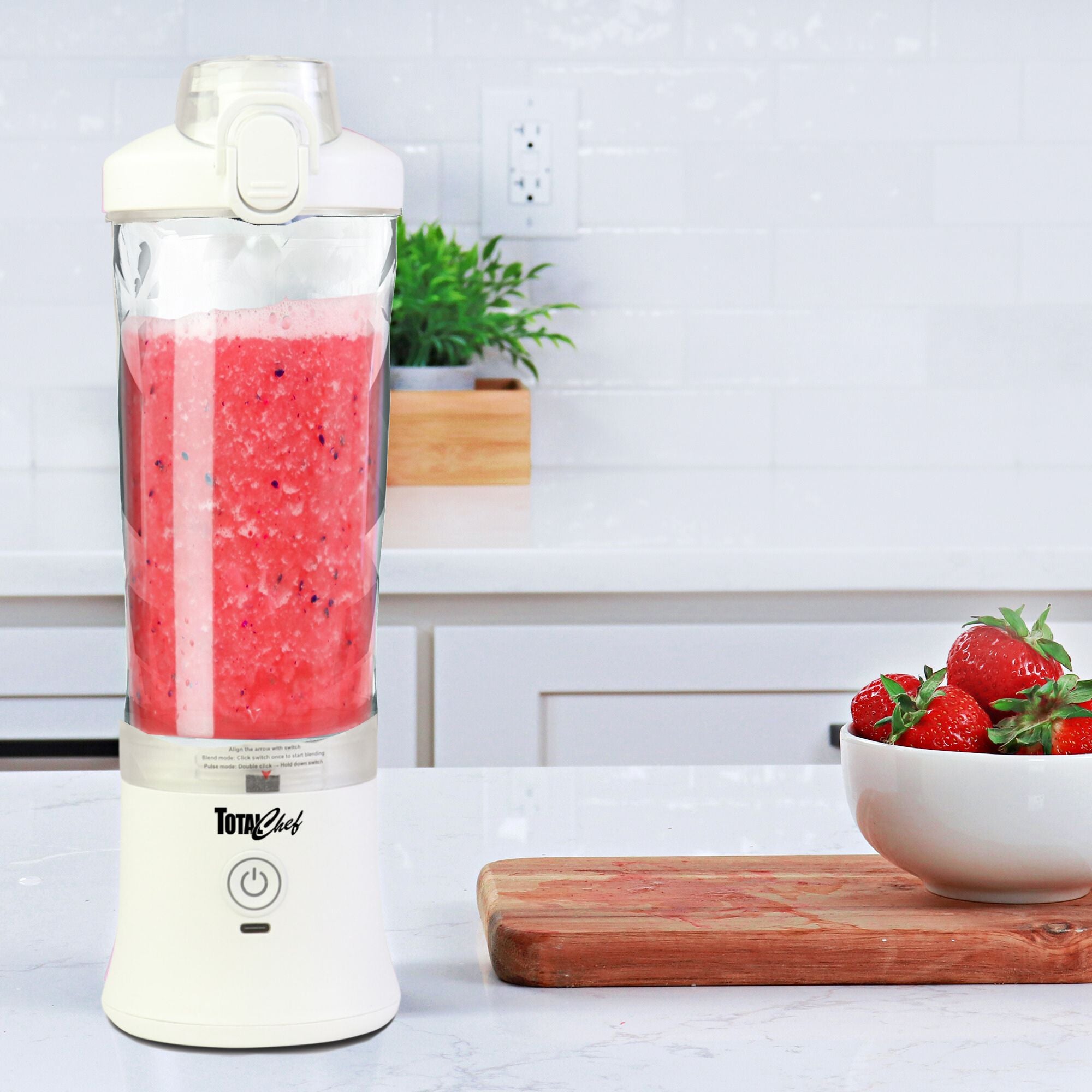 Lifestyle image of the Total Chef portable rechargeable blender filled with bright pink smoothie in a white kitchen with a wooden cutting board and a white bowl of strawberries beside it.